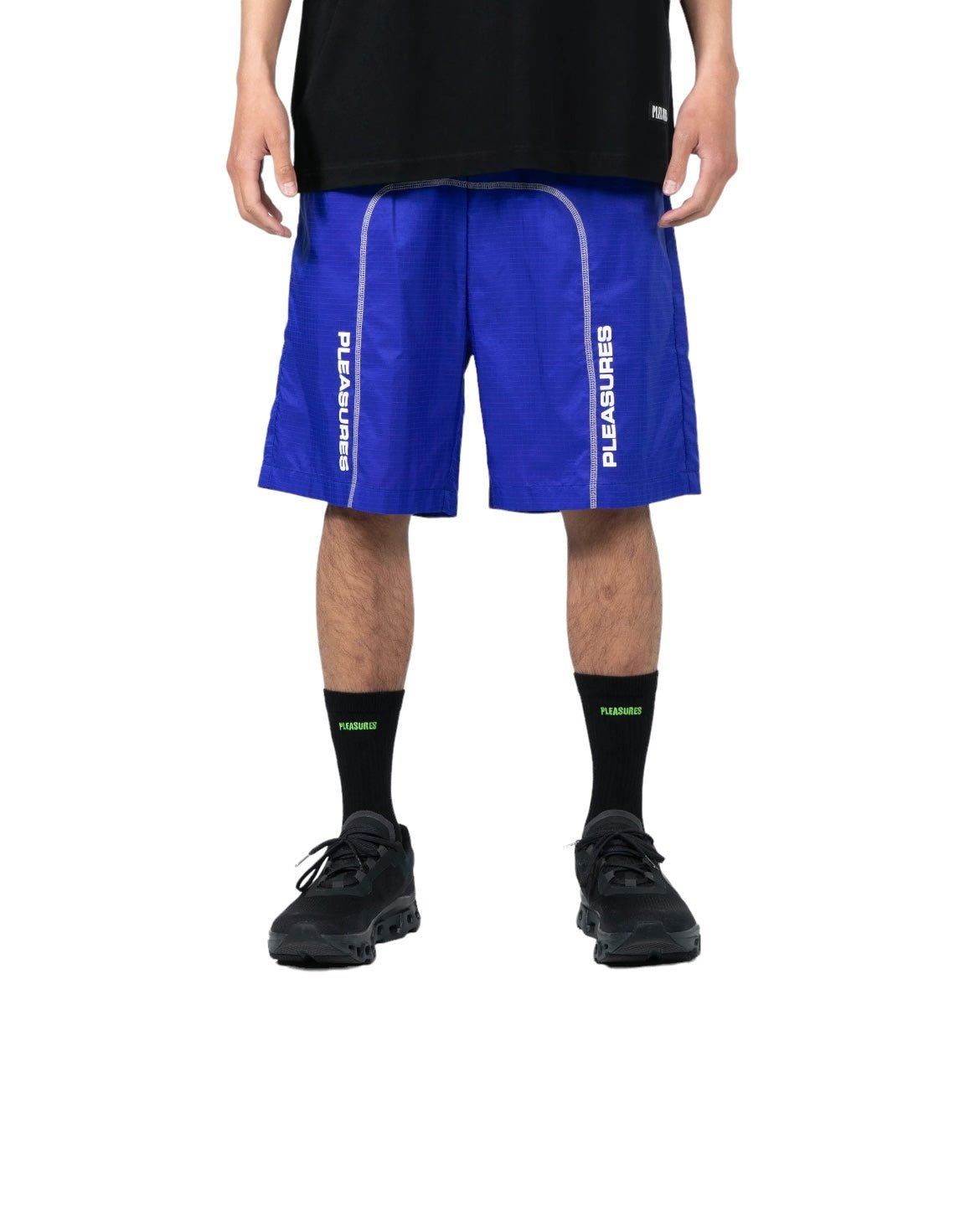 Person wearing a black T-shirt, blue PLEASURES TEMPO ACTIVE SHORTS BLUE by PLEASURES, black socks with neon green detail, and black shoes, standing against a plain white background.