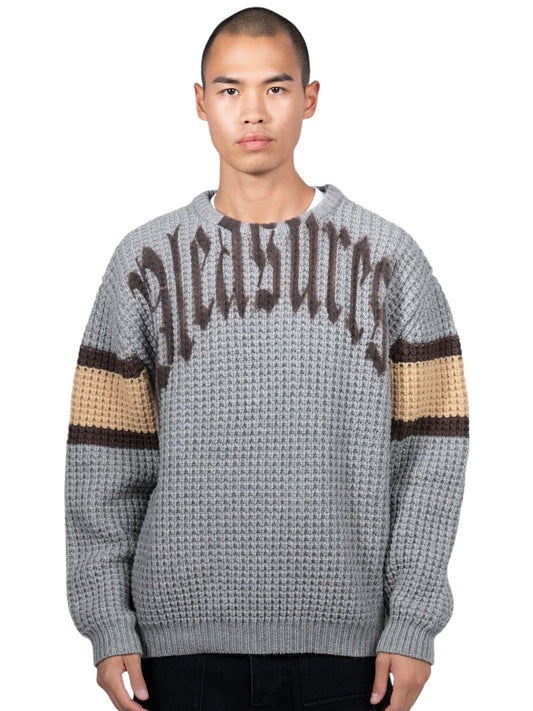 Someone sports the "Pleasures Twitch Chunky Knit Sweat Grey," featuring wool blend and "PLEASURES" embroidery on the front, with chic brown and beige sleeve bands.