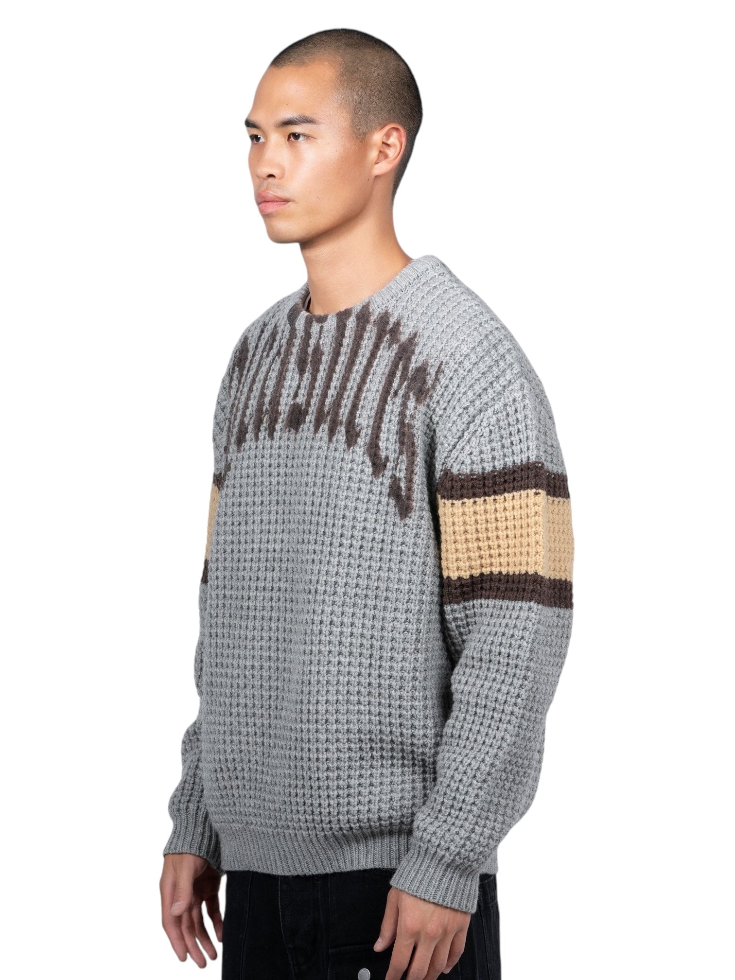 Someone is wearing the Pleasures Twitch Chunky Knit Sweat Grey by PLEASURES, featuring brown and yellow striped sleeves, against a white backdrop.