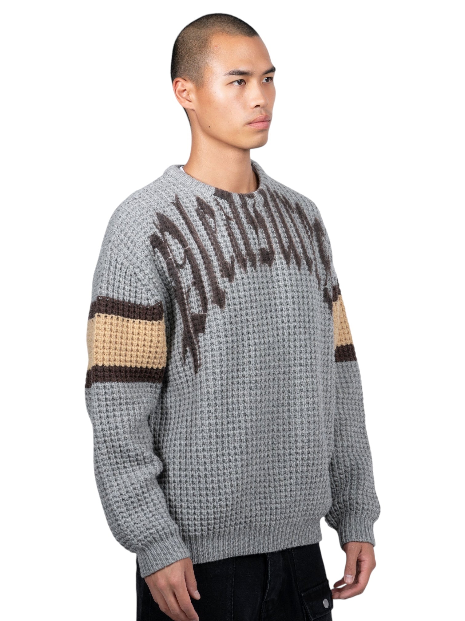 A person wears the Pleasures Twitch Chunky Knit Sweat Grey by PLEASURES, featuring a wool blend with brown and beige stripes, against a white backdrop.