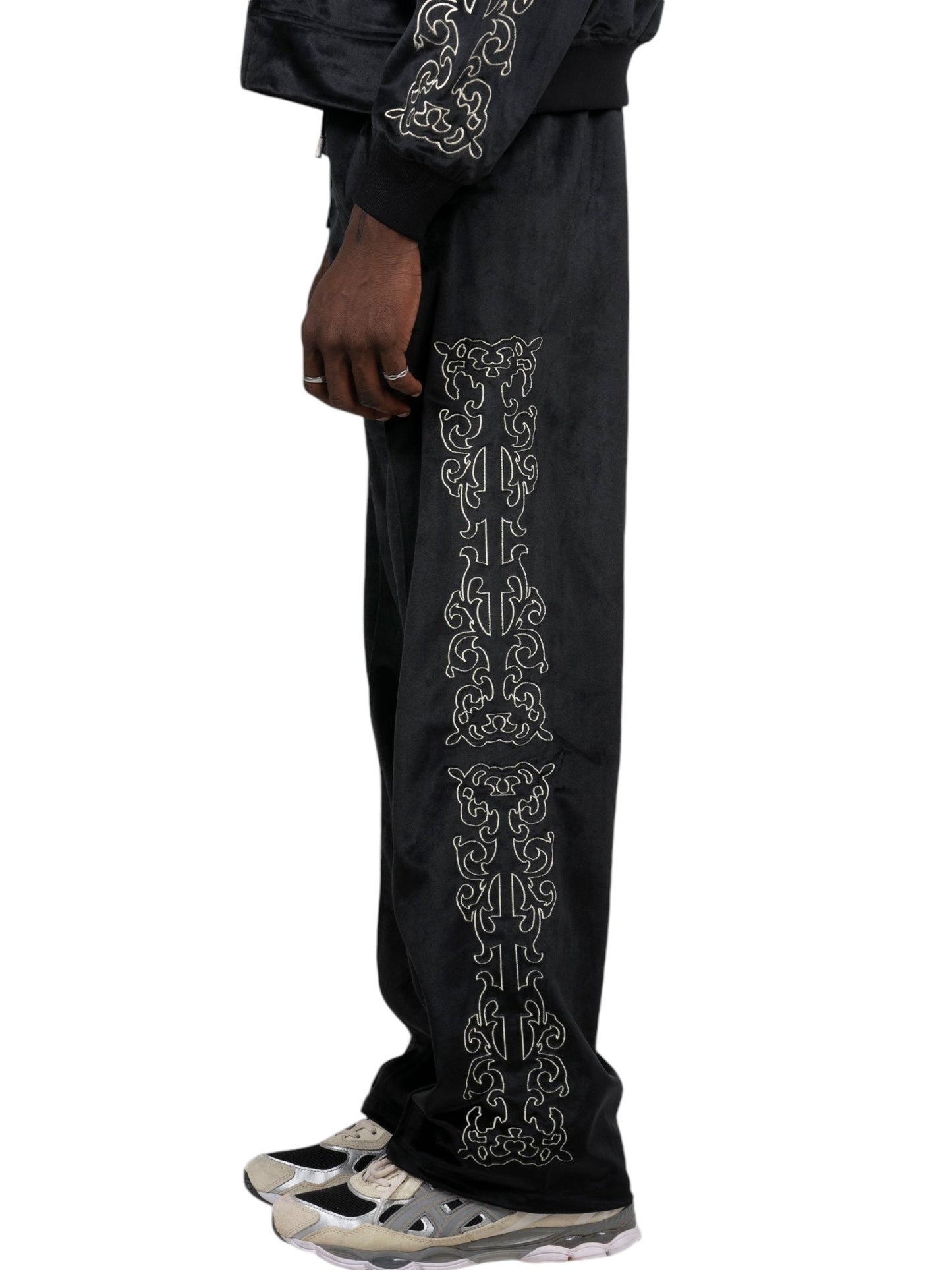 A person styled in Pleasures Unquote Velour Track Pants by PLEASURES, featuring intricate white embroidery along the sides, paired seamlessly with crisp white sneakers.