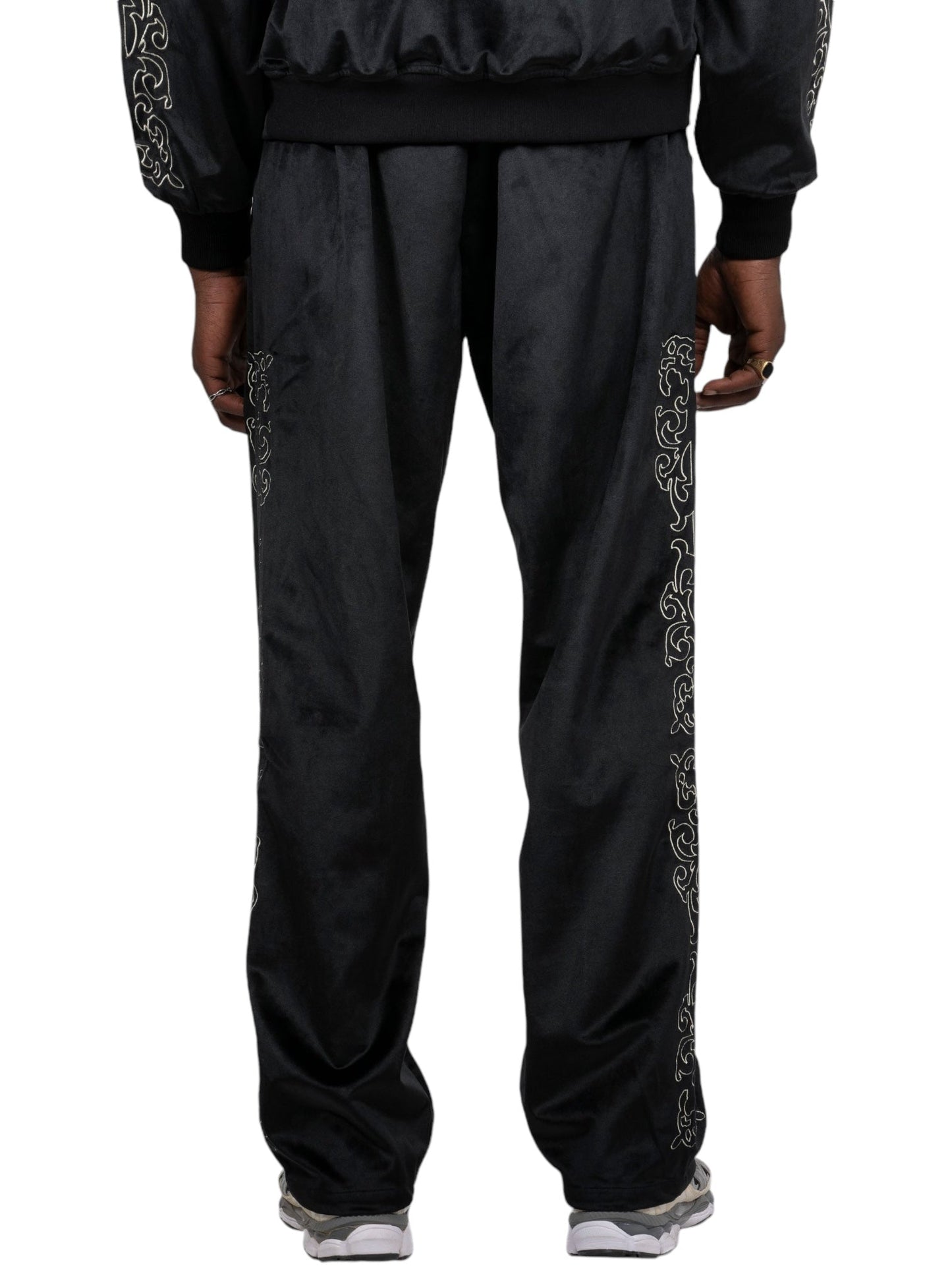 A person wearing Pleasures Unquote Velour Track Pants in black by PLEASURES, featuring ornate embroidery along the sides, stands with their back to the camera. White sneakers peek out at the bottom.