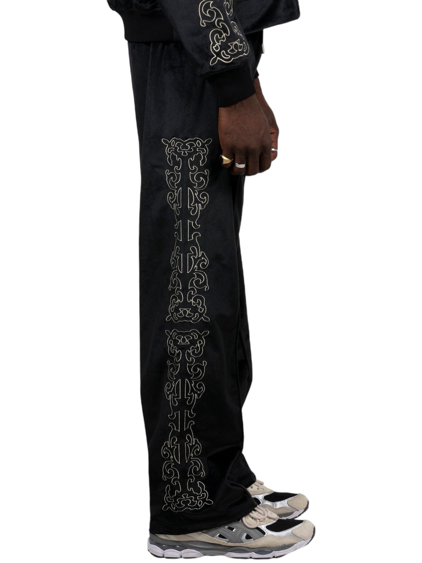 A person wearing Pleasures Unquote Velour Track Pants Black by PLEASURES with ornate side embroidery and sneakers stands against a white background, visible only from the waist down.