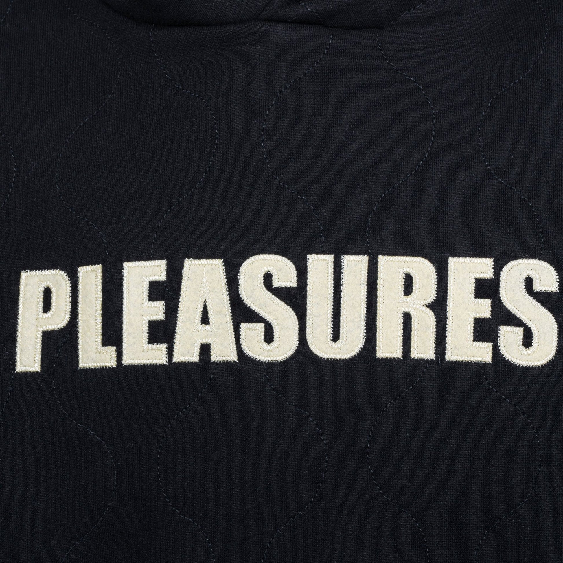 Close-up of the Pleasures Wave Quilted Hoodie Black by PLEASURES, showcasing a unique applique patch with "PLEASURES" embroidered in bold white letters.