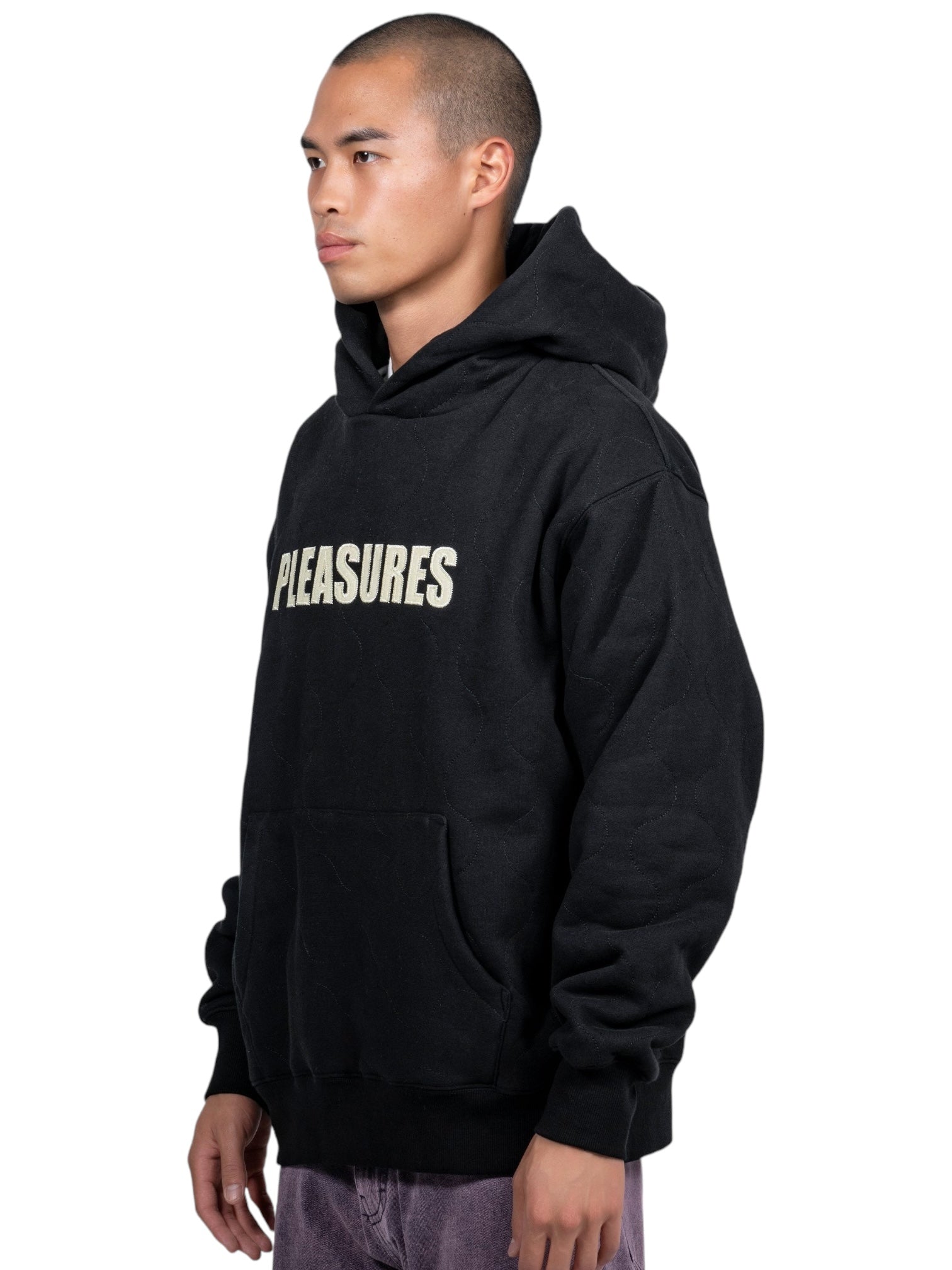 A person wearing the Pleasures Wave Quilted Hoodie Black with "PLEASURES" on the front and a unique applique patch stands against a plain white background.