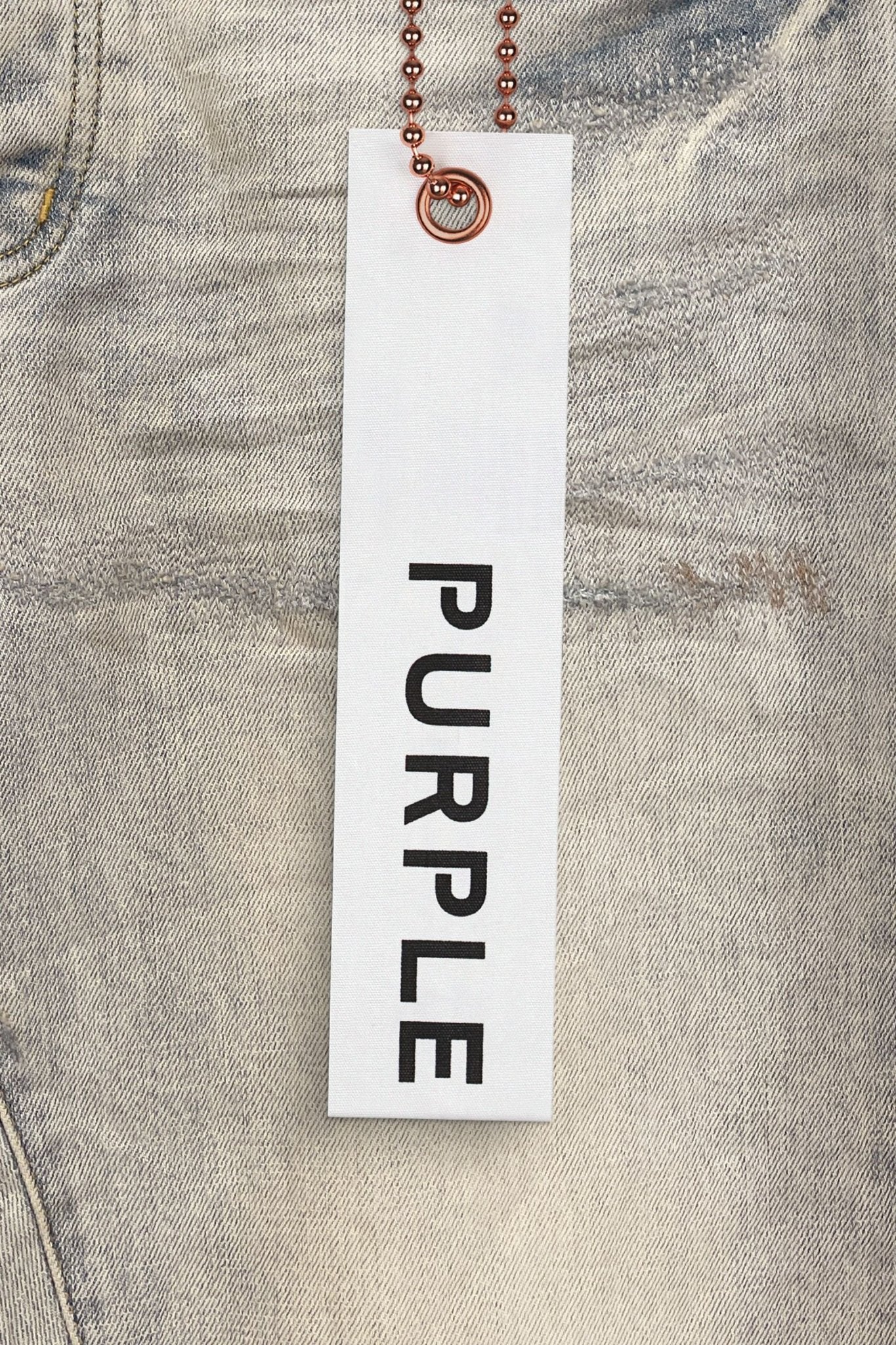 A close-up of Purple Brand Jeans P001-SOR Superlight Oil Repair reveals reinforced belt loops for durability, featuring a white PURPLE BRAND hangtag attached by a ball chain.