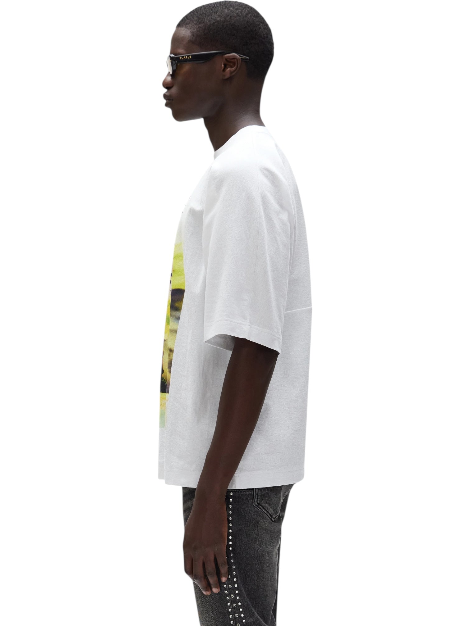 A person stands in profile wearing a PURPLE BRAND M1057-TWFM325 Textured Jersey Top in white, paired with dark studded jeans and sunglasses, set against a plain white background.