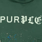 The Purple Brand M4104-HFGH425 Heavyweight Fleece Hoodie in green features an oversized fit, white "PURPLE" crackle print, and blue and beige paint splatters below the text.
