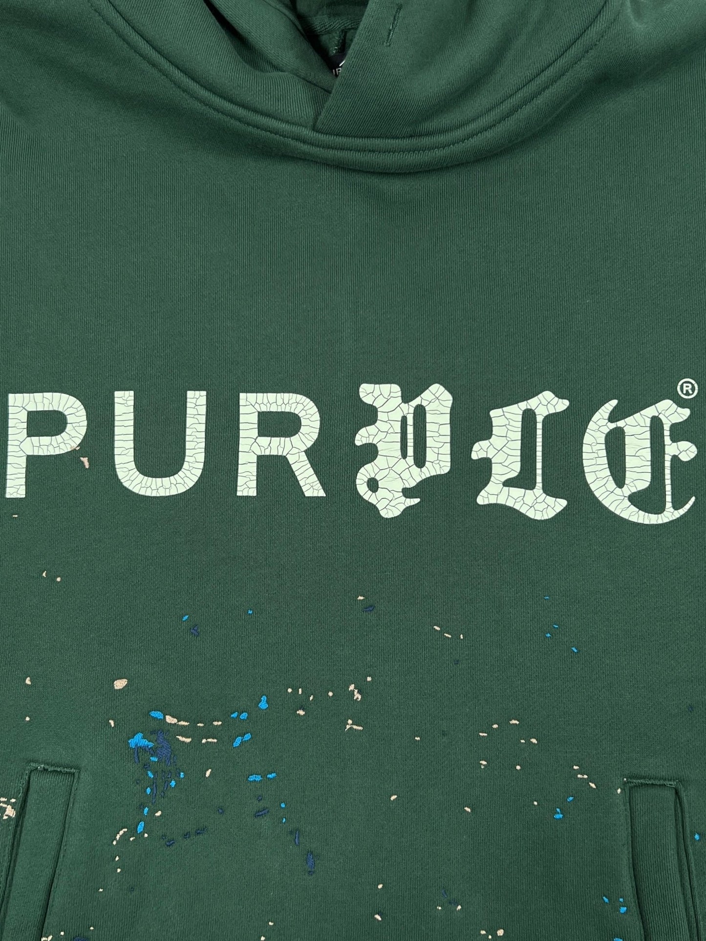 The Purple Brand M4104-HFGH425 Heavyweight Fleece Hoodie in green features an oversized fit, white "PURPLE" crackle print, and blue and beige paint splatters below the text.