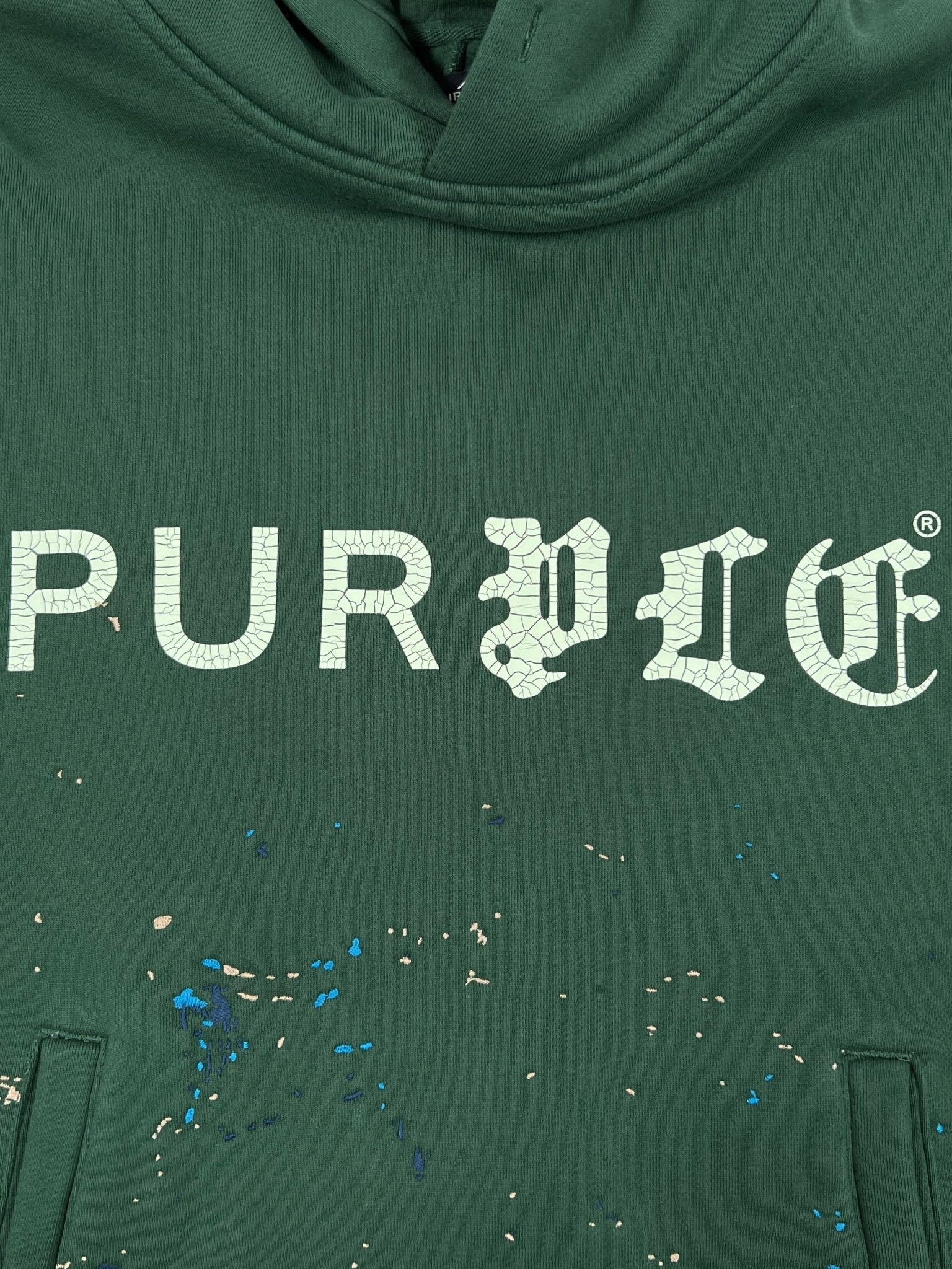 The Purple Brand M4104-HFGH425 Heavyweight Fleece Hoodie in green features an oversized fit, white "PURPLE" crackle print, and blue and beige paint splatters below the text.