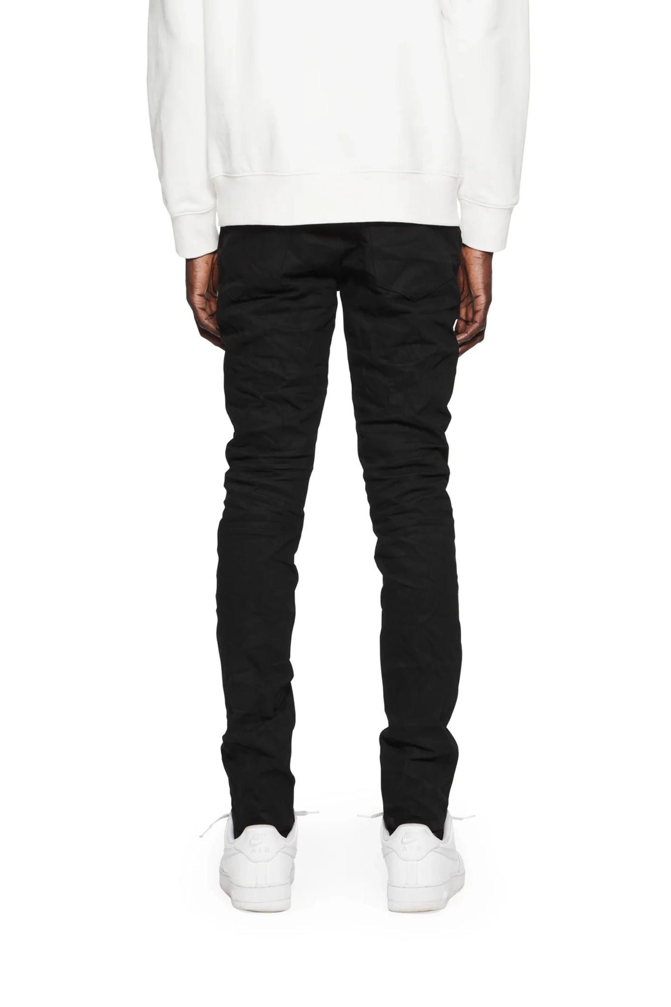 A person in a white sweater and PURPLE BRAND P001-BLKR Black Raw222 jeans, with a dark black wash, stands facing away, paired with white sneakers.