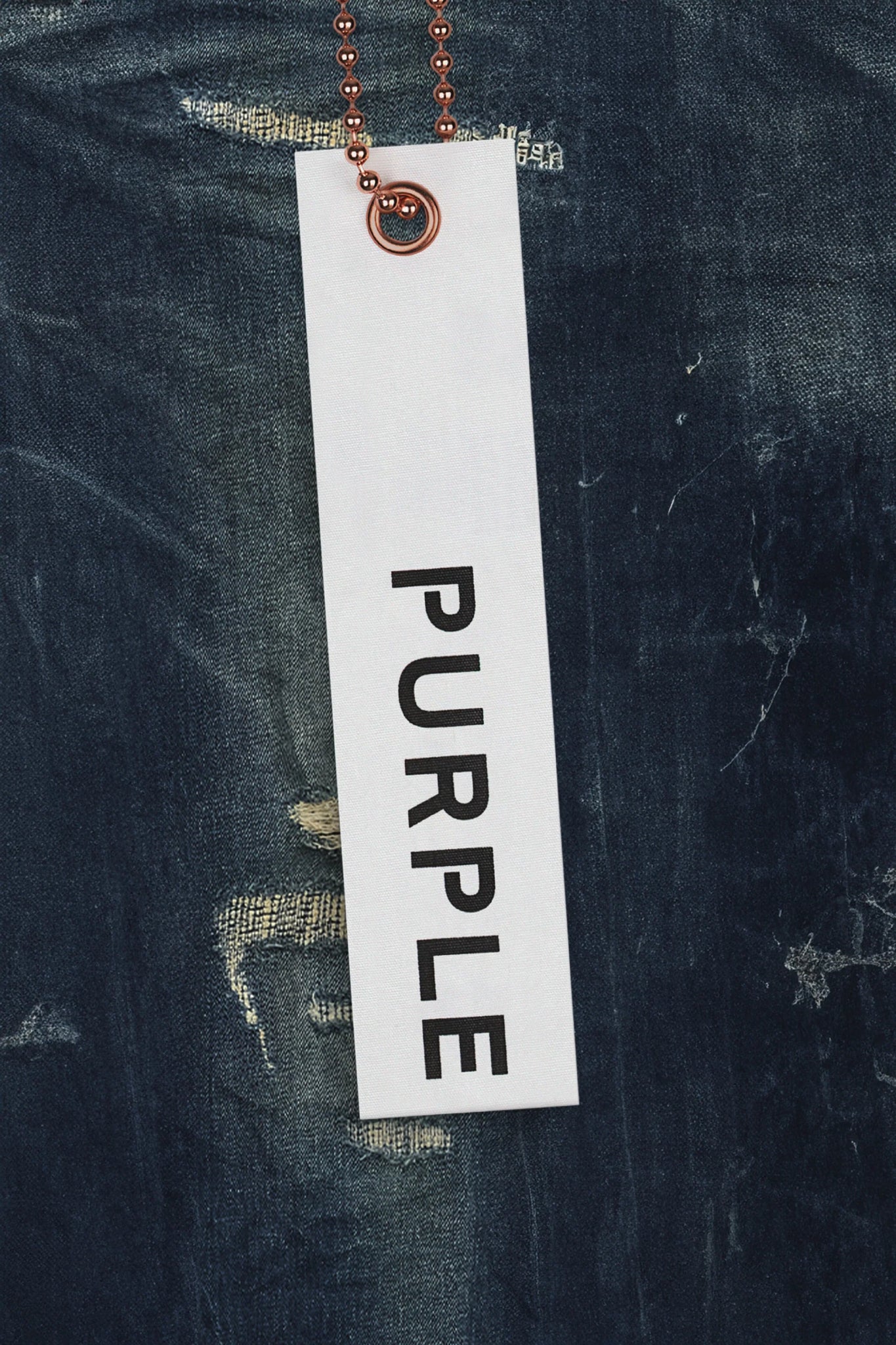 A tag with the word "PURPLE" in black uppercase letters hangs on a copper ball chain against a background of distressed PURPLE BRAND P001-LDDI LIGHT DESTROY DK INDIGO premium stretch denim fabric.