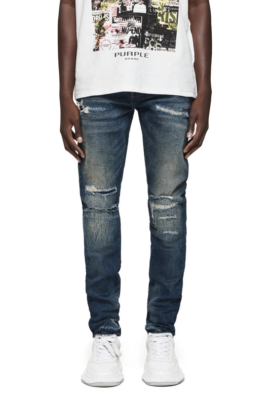 A person wearing a white printed shirt, Purple Brand P001-LDDI Light Destroy Dk Indigo jeans from PURPLE BRAND made with premium stretch denim, and white sneakers stands against a white background.