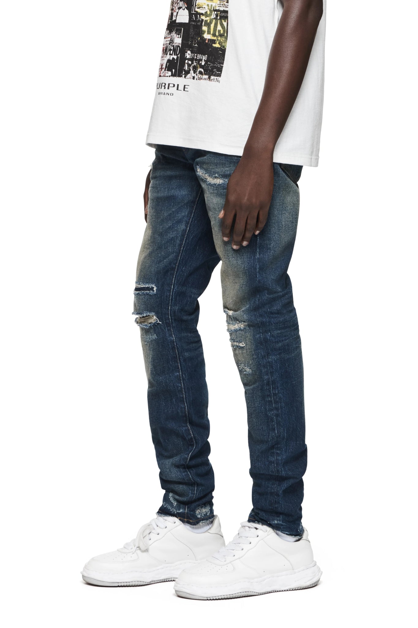 A person wearing PURPLE BRAND P001-LDDI LIGHT DESTROY DK INDIGO low rise skinny jeans with distressed patches, a white graphic t-shirt, and white sneakers stands against a white background.