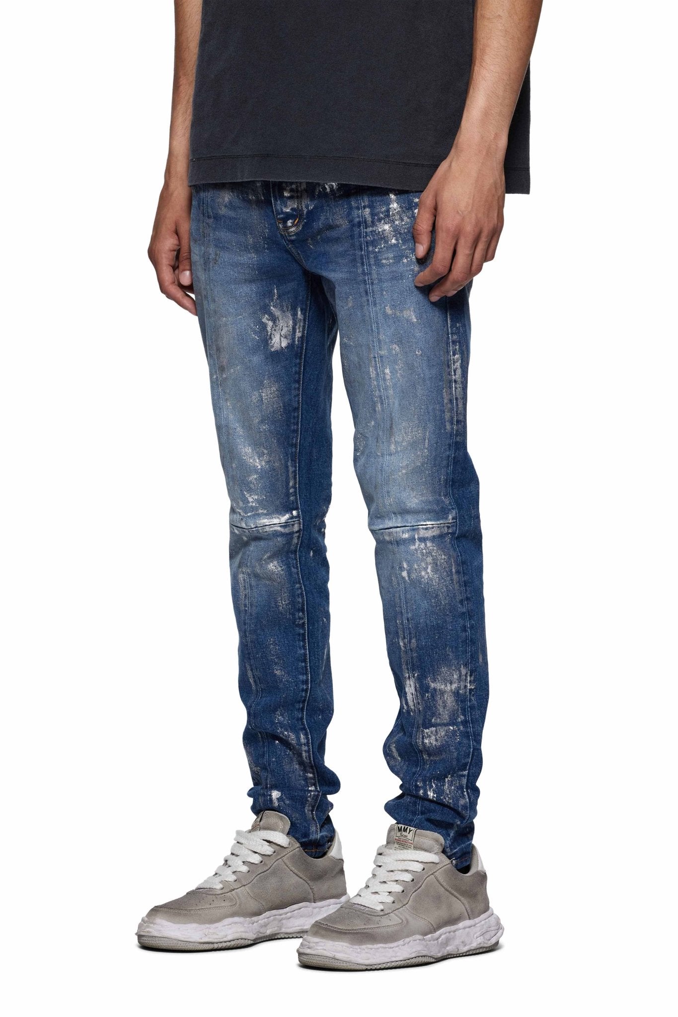 A person wearing PURPLE BRAND's P001-SAFC Angel Silver Foil Cross Indigo low-rise skinny jeans with unique washes, a black shirt, and gray sneakers stands against a plain white background.