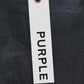 A Metallic chain suspends a white tag reading "PURPLE" in black text against dark fabric, evoking the premium stretch denim of Purple Brand's P001-SCBI Skinny Black Coated Indigo jeans.