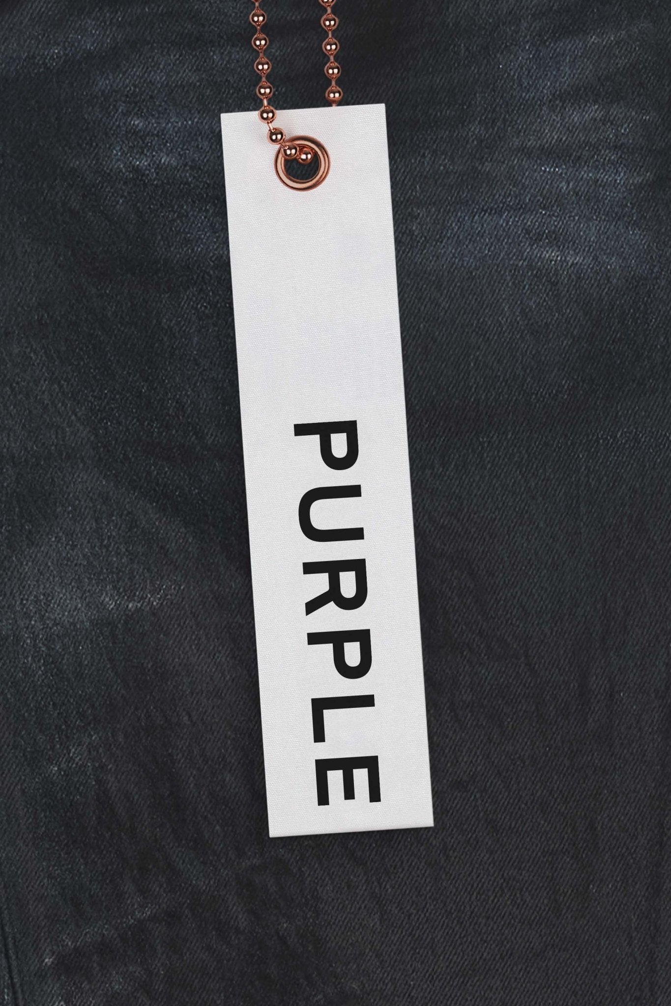 A Metallic chain suspends a white tag reading "PURPLE" in black text against dark fabric, evoking the premium stretch denim of Purple Brand's P001-SCBI Skinny Black Coated Indigo jeans.
