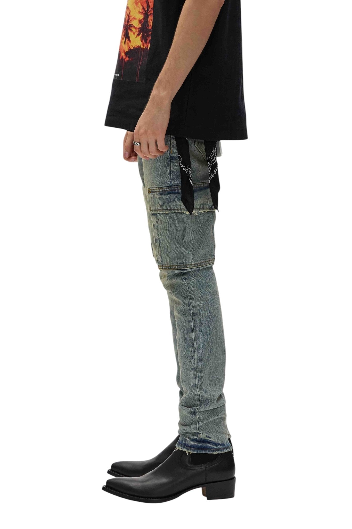 A person in a black shirt and PURPLE BRAND's P001-SCPL Skinny Cargo Pumice Lt Indigo jeans with patchwork stands sideways. Black pointed-toe boots complete the look, while a black bandana hangs casually from a back pocket.