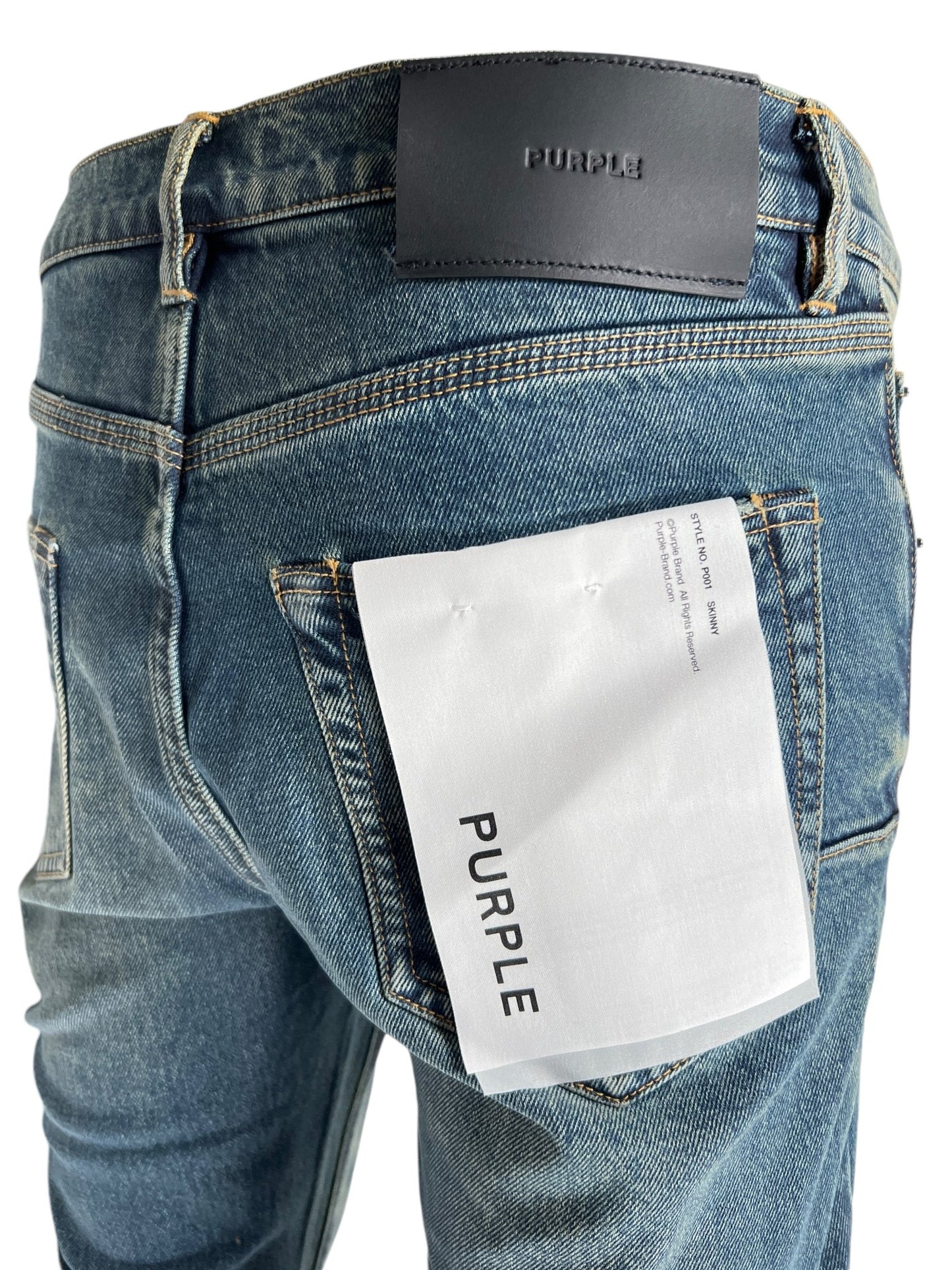 Close-up of the back of a person wearing PURPLE BRAND P001-SDTE SKINNY COATED DK TEMPEST DK INDIGO jeans with a brand label "PURPLE" above the right pocket and a large white tag tucked into the pocket, highlighting their skinny fit.