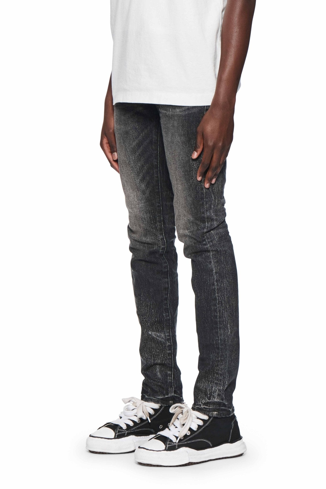 A person wearing a white shirt, PURPLE BRAND P001-SIWC Skinny Crackle Coated Black jeans, and black and white sneakers standing against a plain white background.