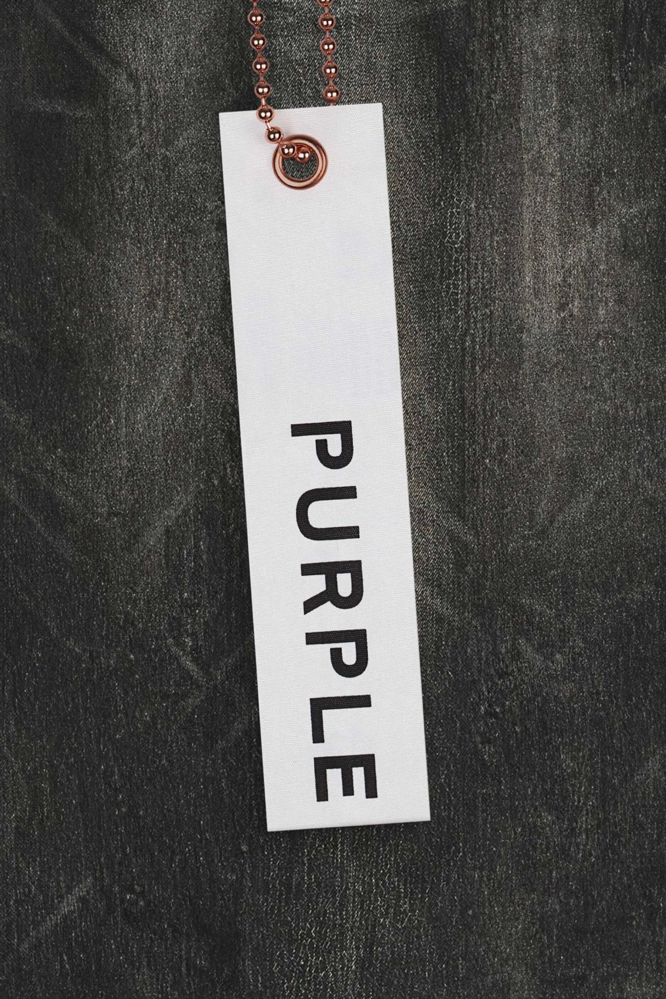 A vertical white tag with the product name "PURPLE BRAND P001-SIWC SKINNY CRACKLE COATED BLACK" written in black font is attached to a metal ball chain, evoking a vintage look against a dark textured background.