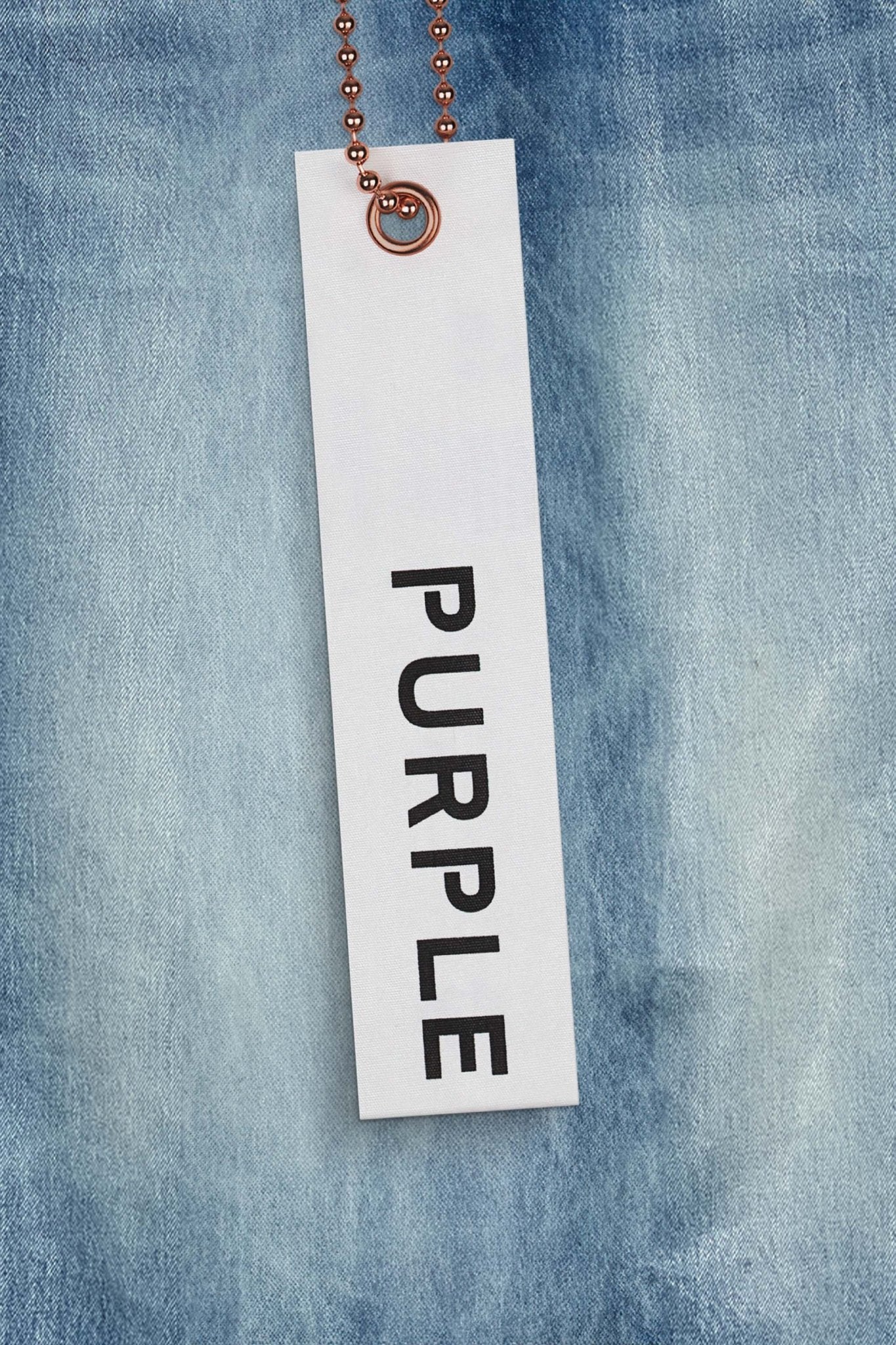 A tag from PURPLE BRAND P001-SKCI SKINNY COATED LT INDIGO, featuring the word "PURPLE" in black letters on a white background and attached to a copper ball chain, lies on a backdrop of premium stretch denim fabric.