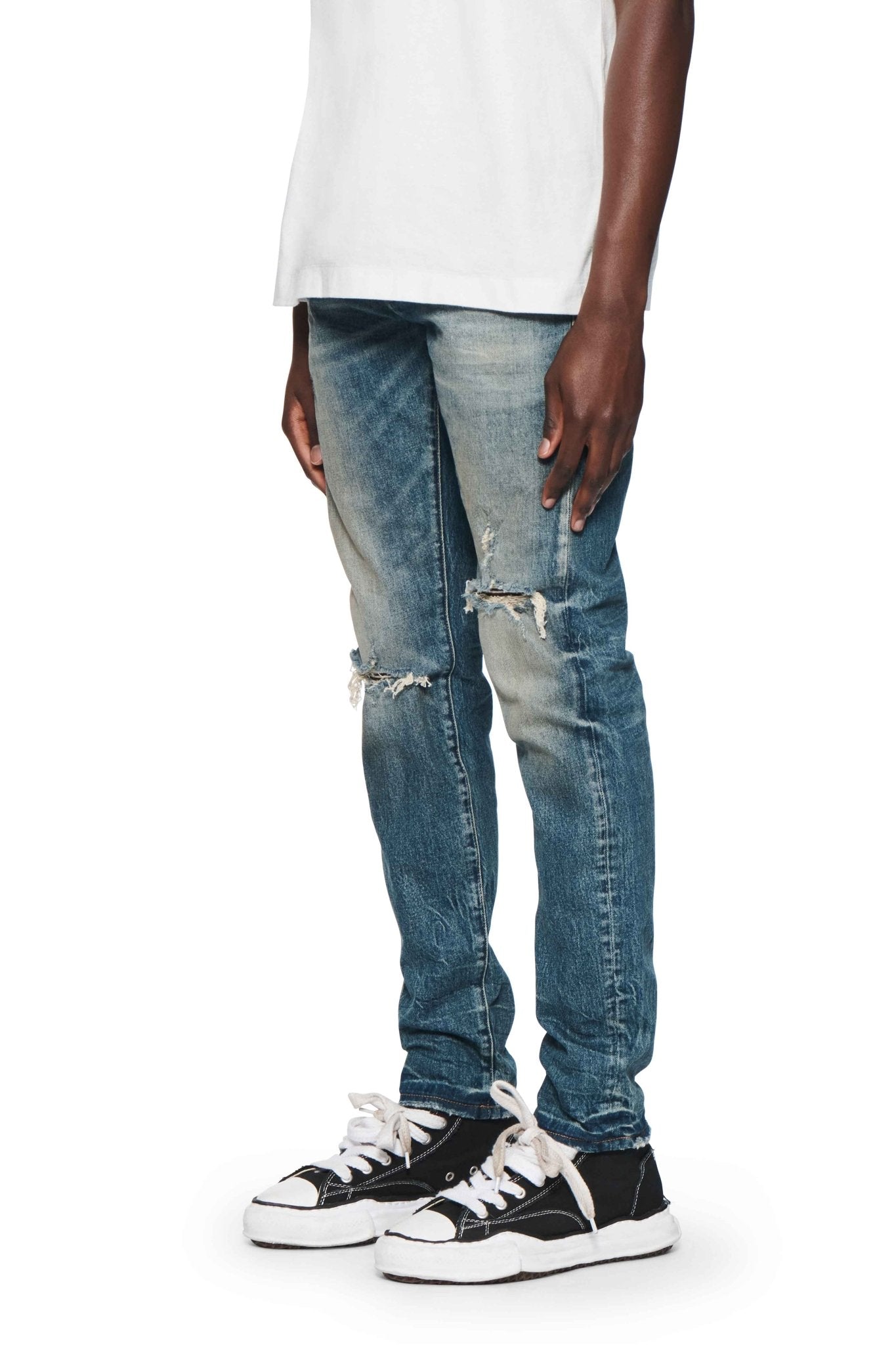 Man wearing a white shirt, PURPLE BRAND P001-SNFD SKINNY 9 TO 5 DK INDIGO distressed premium stretch denim blue jeans for Fall Winter 2024 with tears, and black and white sneakers.