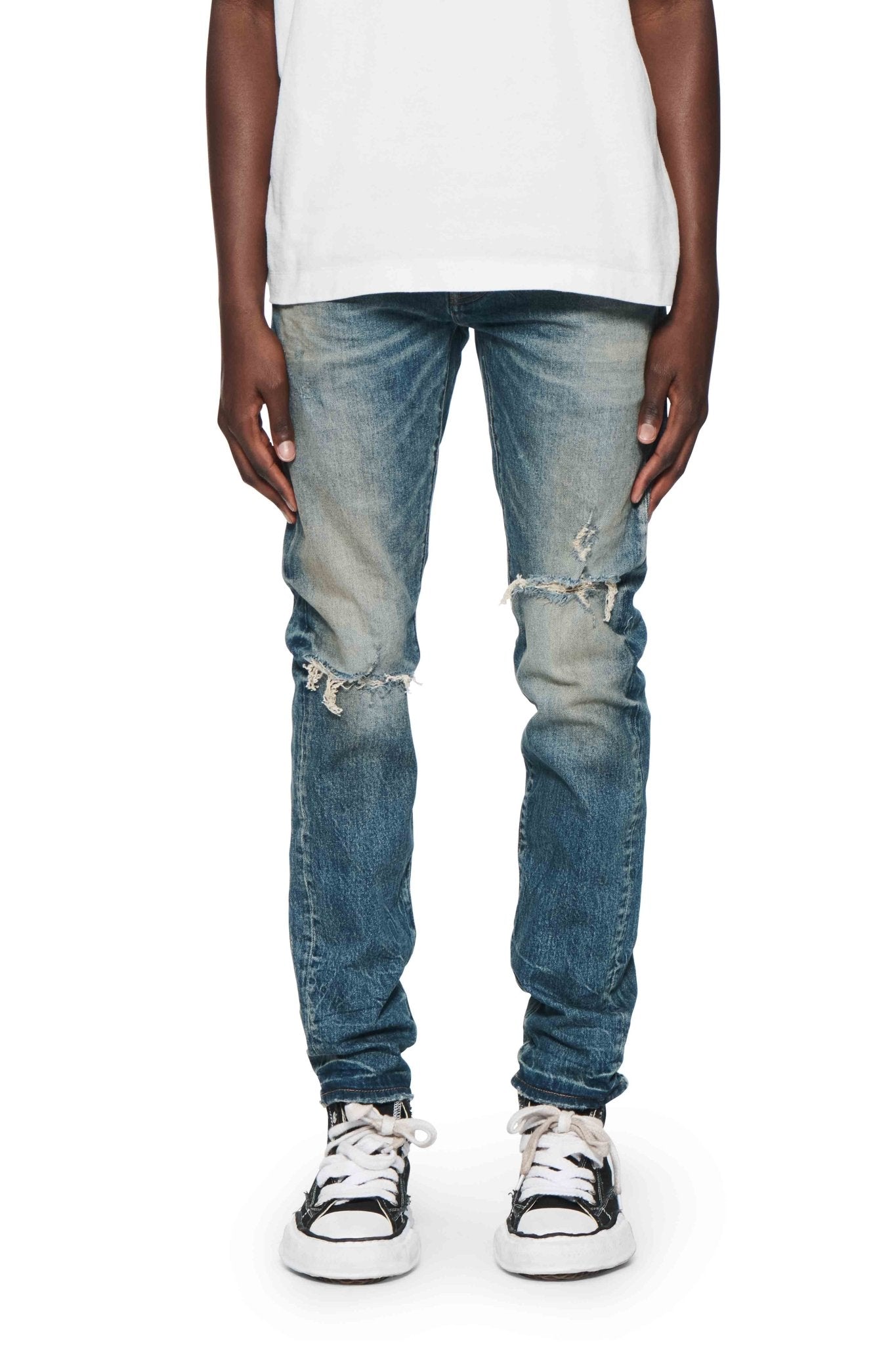 A person is wearing the Purple Brand P001-SNFD Skinny 9 To 5 DK Indigo denim with white sneakers, standing facing forward with their hands at their sides. A low-rise white shirt is partially visible.