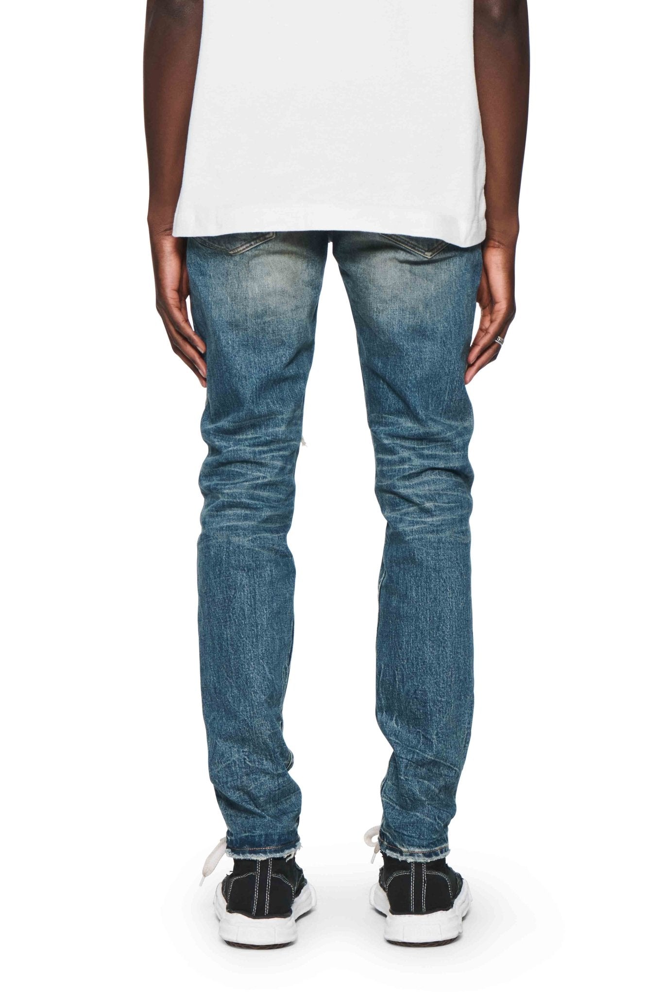 Back view of a person wearing a white shirt, PURPLE BRAND P001-SNFD SKINNY 9 TO 5 DK INDIGO jeans, and black sneakers with white soles.