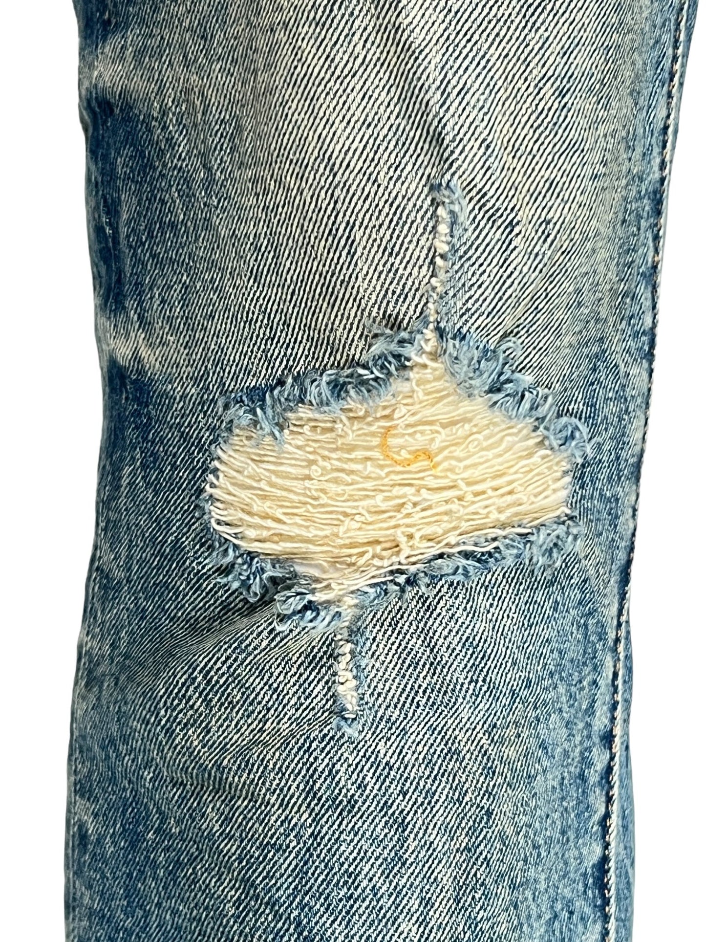 Close-up of a worn and ripped knee on the dark indigo denim, revealing white fabric underneath. The tear is frayed along the edges, showcasing the rugged appeal of PURPLE BRAND P001-SNFD SKINNY 9 TO 5 DK INDIGO jeans crafted from premium stretch denim.