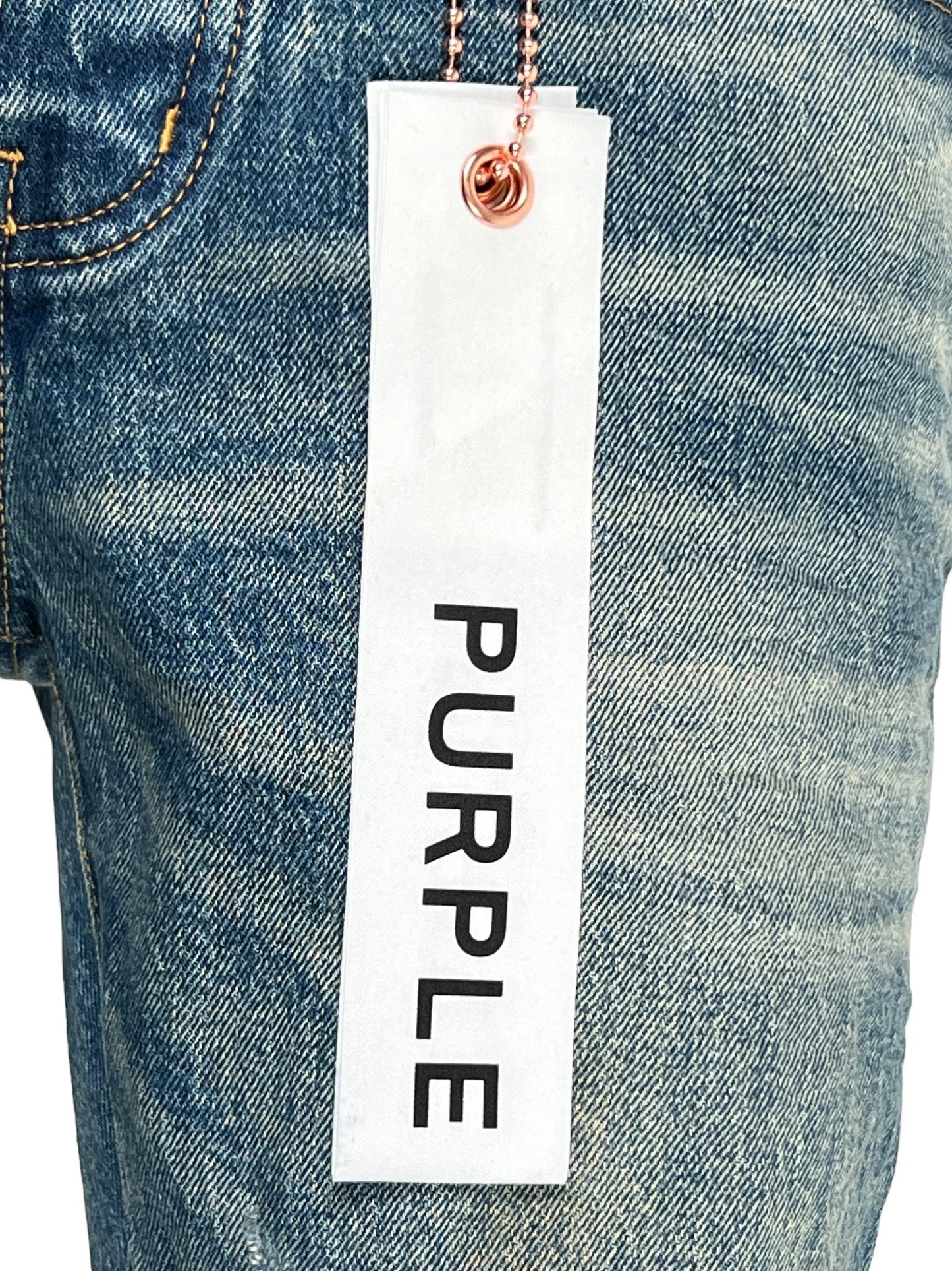 Close-up of PURPLE BRAND P001-SNFD SKINNY 9 TO 5 DK INDIGO jeans in a dark indigo wash with a white tag displaying the word "PURPLE" in black text, hanging from a copper ball chain.