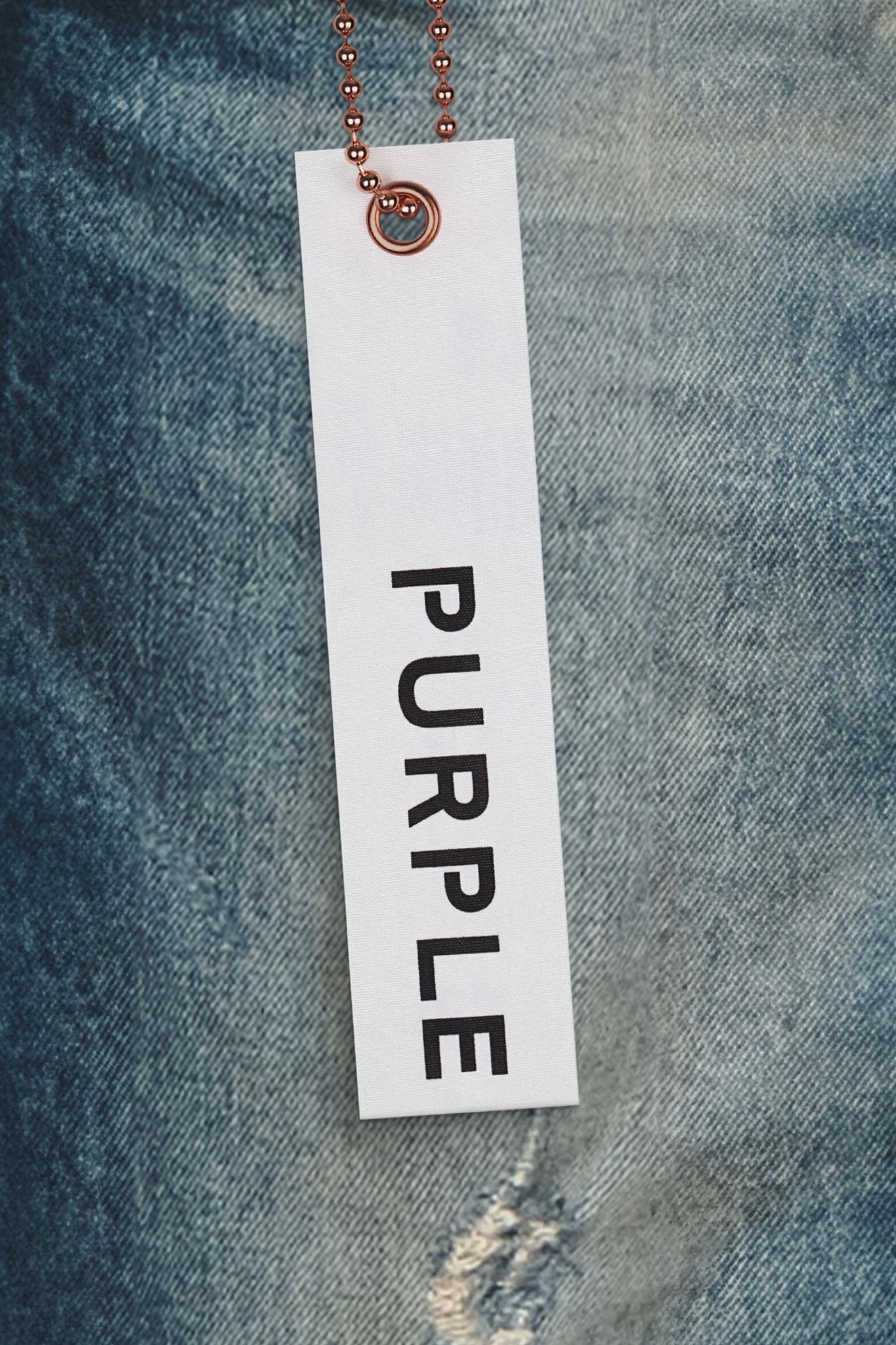 A white tag labeled "PURPLE BRAND" hangs against the blue premium stretch denim, capturing the allure of the chic Purple Brand P001-SNFD Skinny 9 To 5 DK Indigo low rise skinny jeans.