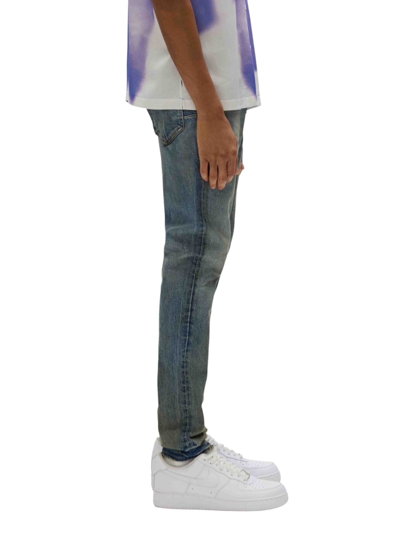 The side profile captures a person wearing a white and blue patterned shirt against a plain background, styled with PURPLE BRAND's P001-SYCB Skinny Cracked Bronze Denim from the Spring/Summer 2025 White Label collection, and paired with white sneakers.