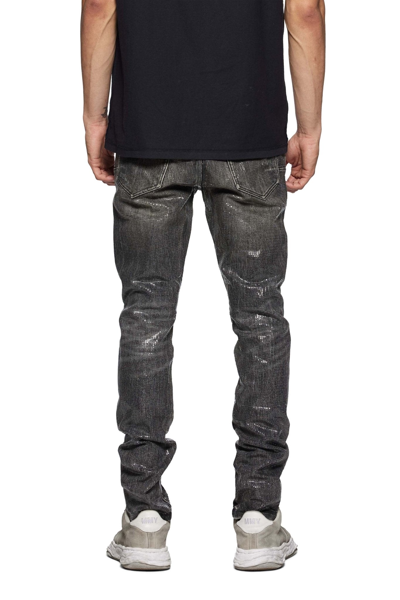 A person stands with their back to the camera, showcasing the Purple Brand P001-TOBL Tiled Overlay Black shirt, shiny gray low-rise skinny jeans made from premium stretch denim, and white sneakers.