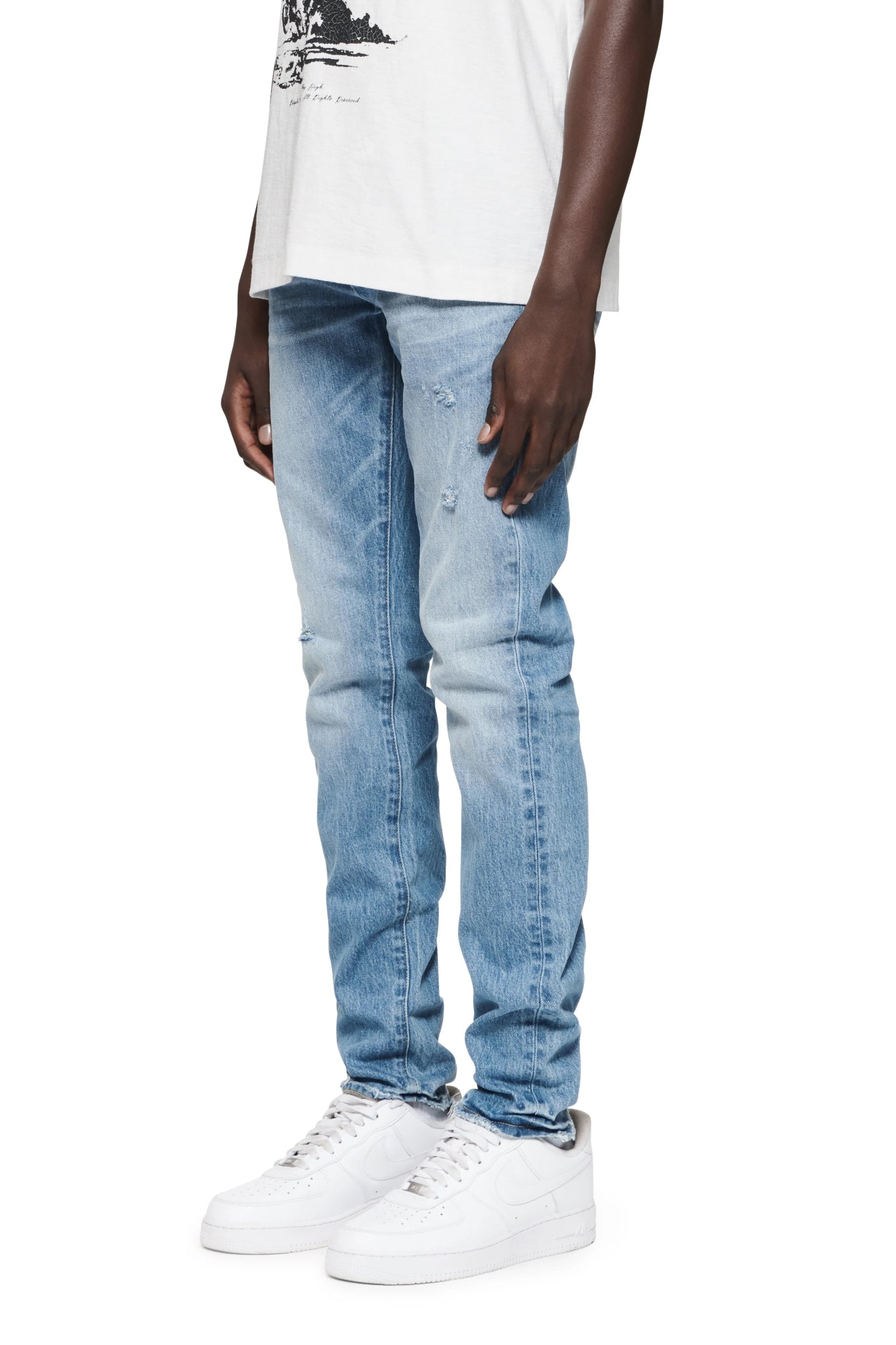 A person wearing a white t-shirt, PURPLE BRAND P001-VACI VINTAGE ABRASIONS LT INDIGO jeans, and white sneakers stands against a plain background.