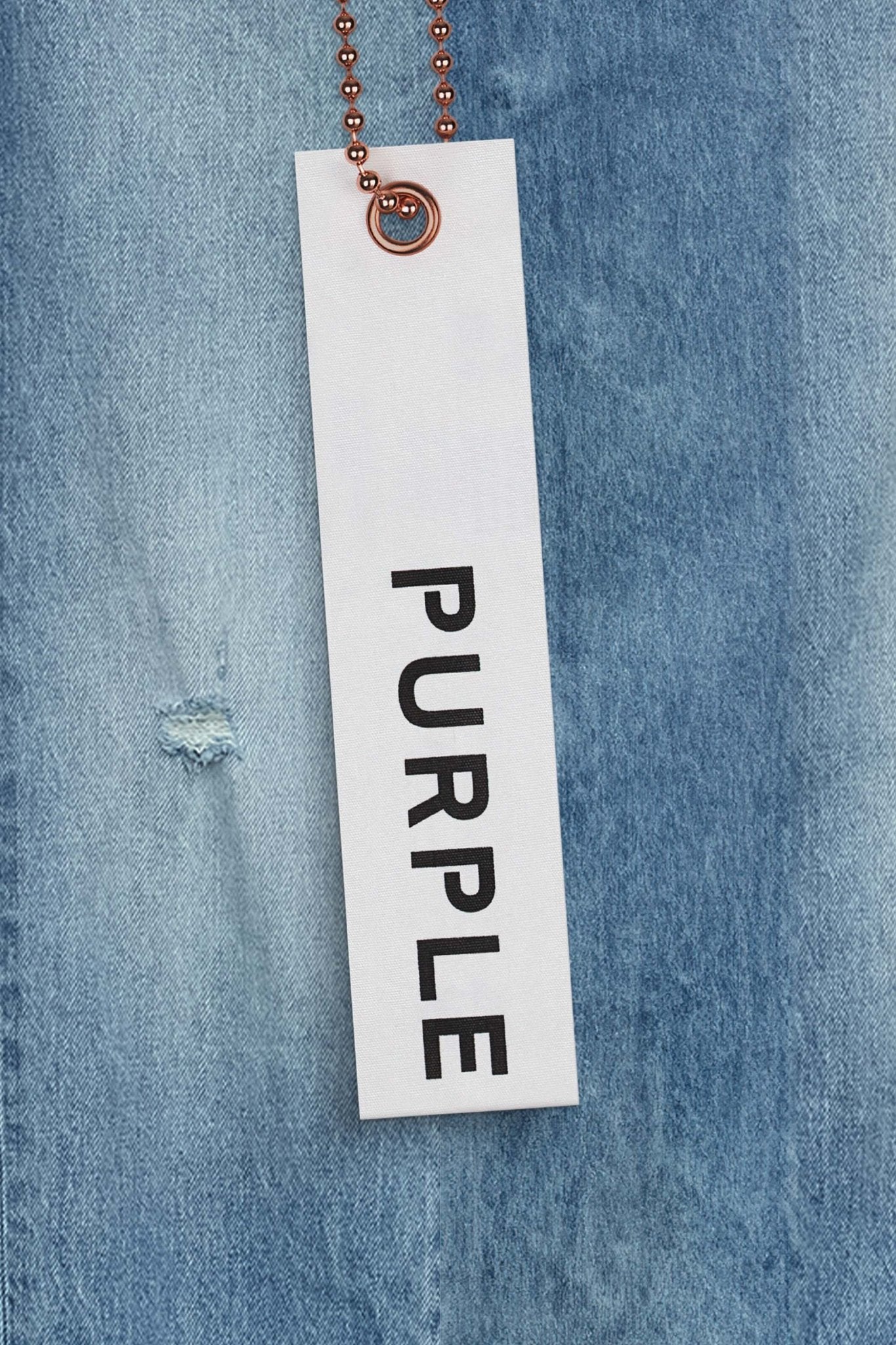 A white tag with the word "PURPLE" in black text is attached to a ball chain and placed against the background of PURPLE BRAND P001-VACI Vintage Abrasions Lt Indigo premium stretch denim, giving it a sleek yet vintage look.