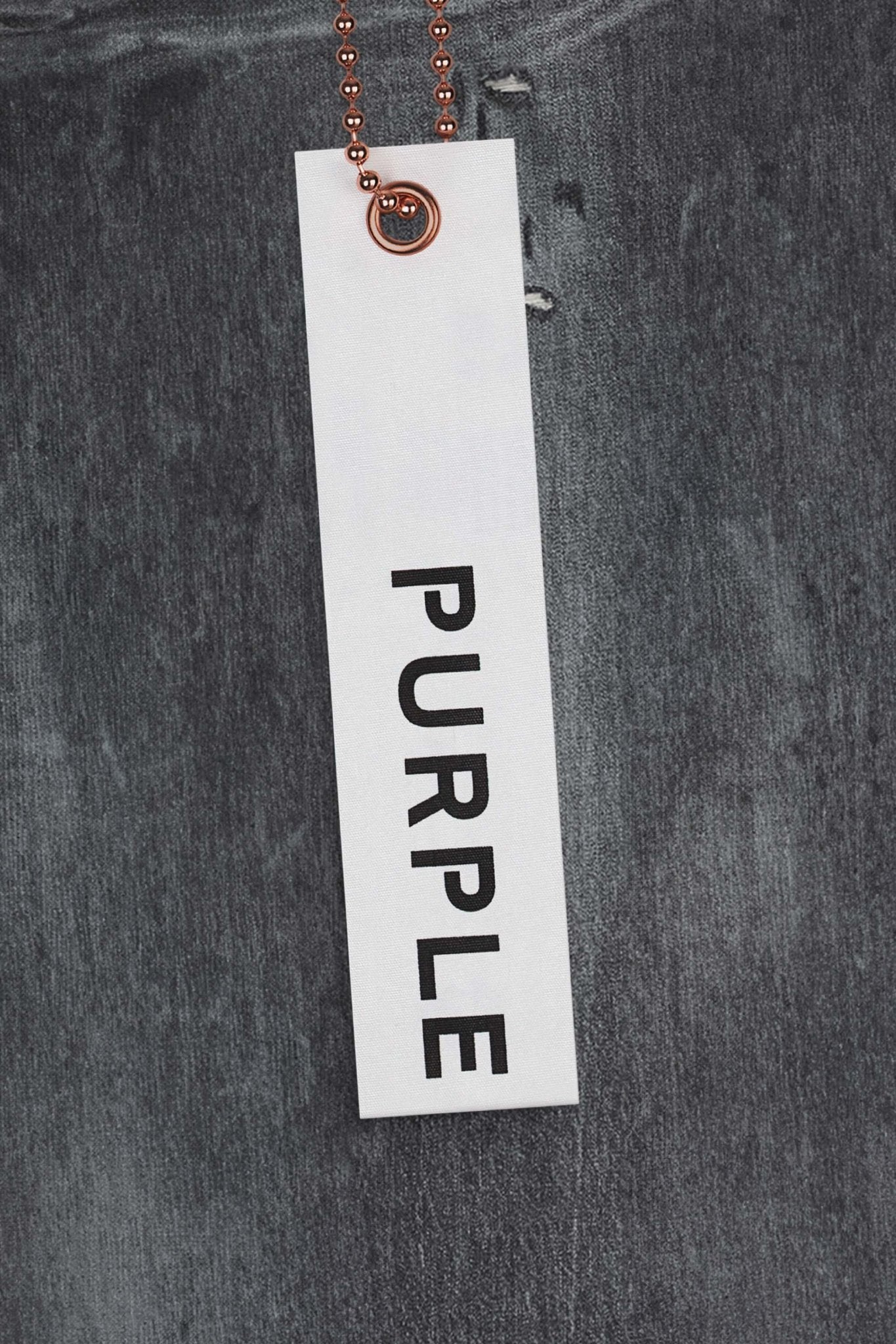 A white tag with the word "PURPLE" written in black, attached to a piece of gray fabric, possibly the PURPLE BRAND P001-VAMG VINTAGE ABRASIONS GREY low rise skinny jeans in premium stretch denim, by a small bronze-colored ball chain.