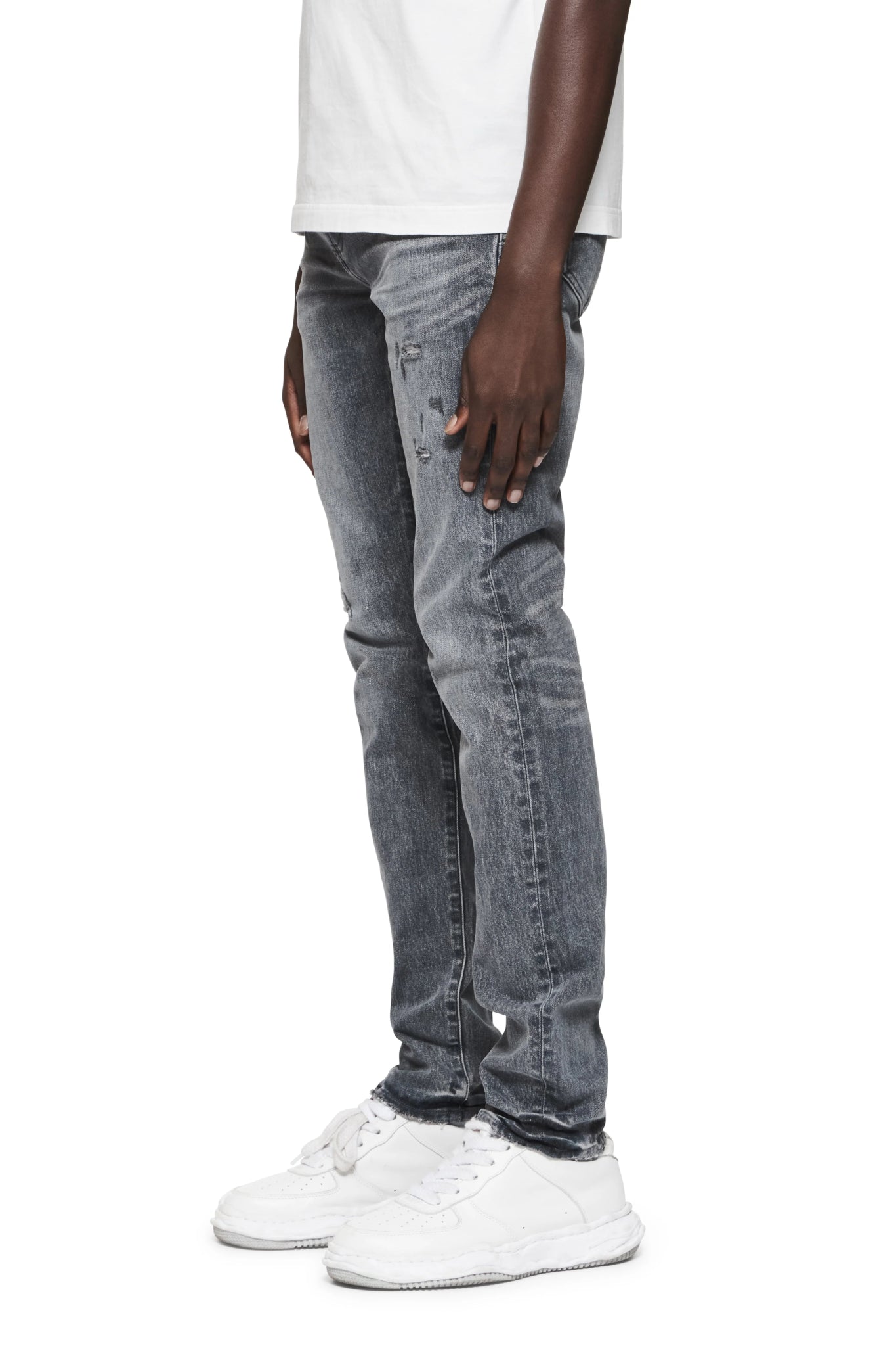 A person wearing a white t-shirt and distressed gray jeans stands with white sneakers visible, showcasing the vintage look of PURPLE BRAND's P001-VAMG Vintage Abrasions Grey premium stretch denim.