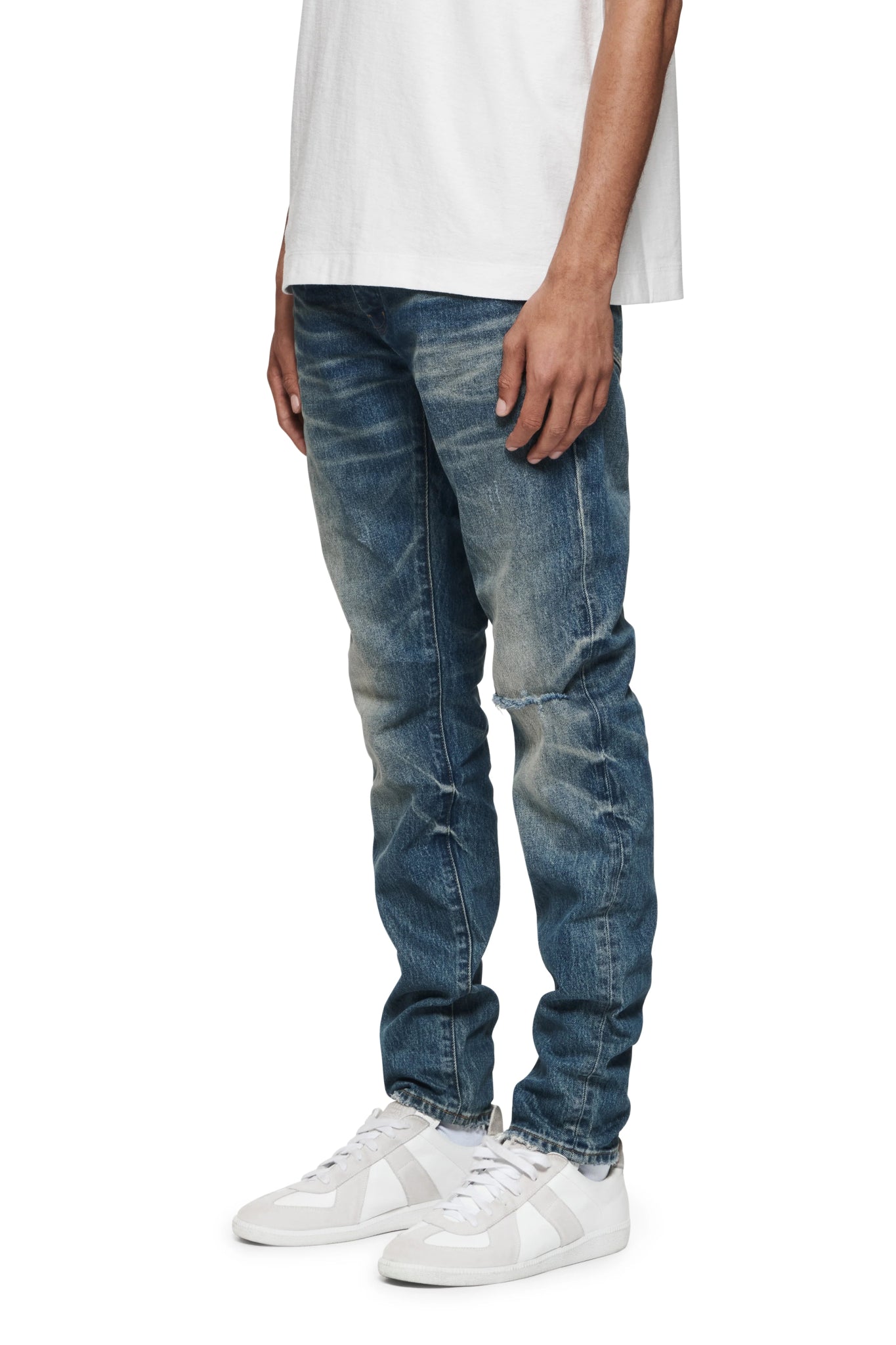 A person wearing a white T-shirt, white sneakers, and PURPLE BRAND P001-VKMD VINTAGE KNEE BLOWOUT MID INDIGO low rise skinny jeans made from premium stretch denim is standing against a plain background. The photo is cropped to show only the lower half of the body, highlighting the handcrafted unique washes of the PURPLE BRAND jeans.