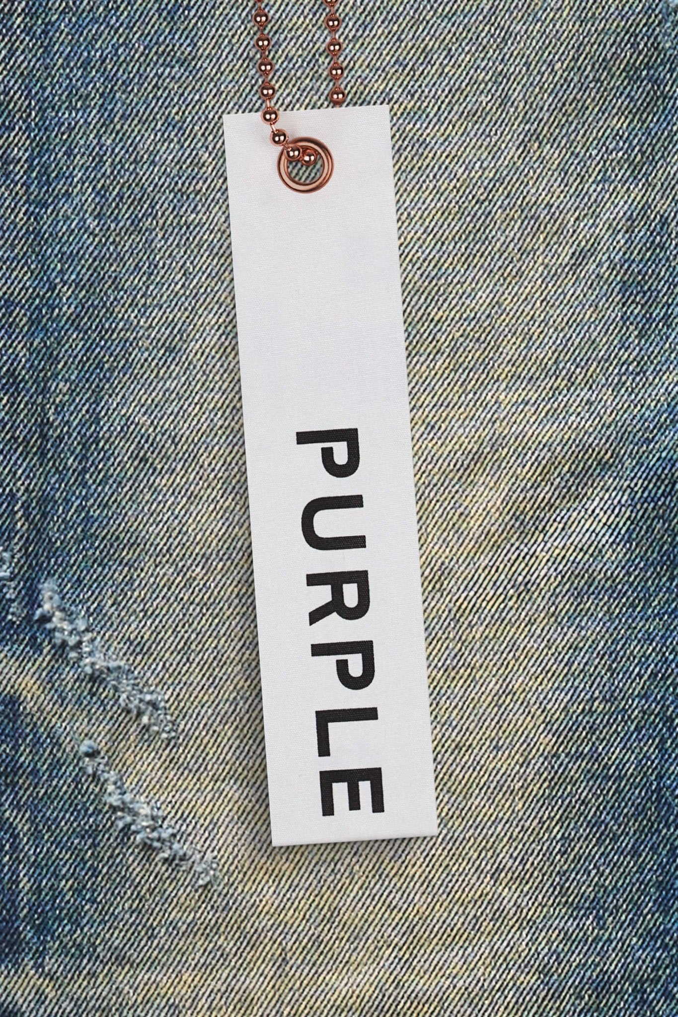 A white rectangular tag with the word "PURPLE" in black letters is attached to a copper-colored ball chain, lying on PURPLE BRAND P001-VKMD VINTAGE KNEE BLOWOUT MID INDIGO premium stretch denim fabric.