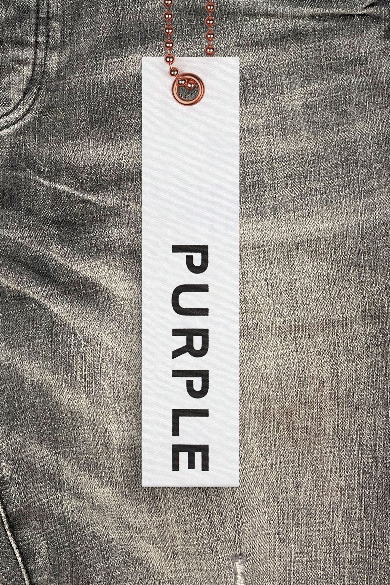 A white tag with "PURPLE" in black hangs by a chain against a pair of PURPLE BRAND P002-VWBO Vintage Worn Blowout Black jeans, crafted from premium stretch denim.
