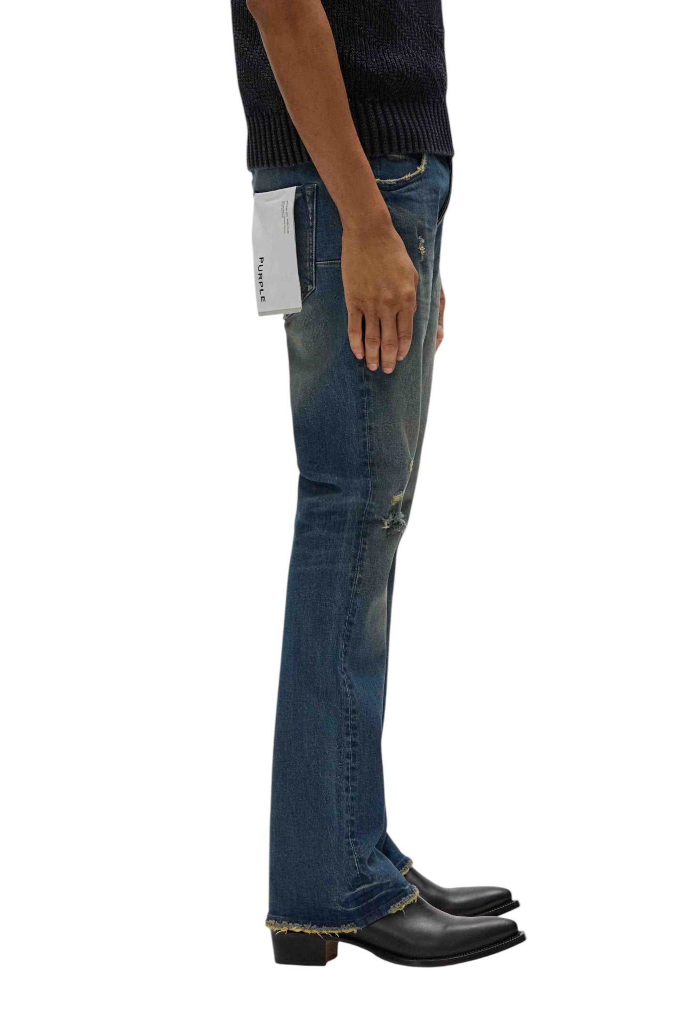 A person stands in profile, wearing a dark short-sleeve top paired with PURPLE BRAND's P004-ALDF Alloy Distressed Flare Indigo jeans, featuring a white pocket label. Black pointed shoes complete the look.