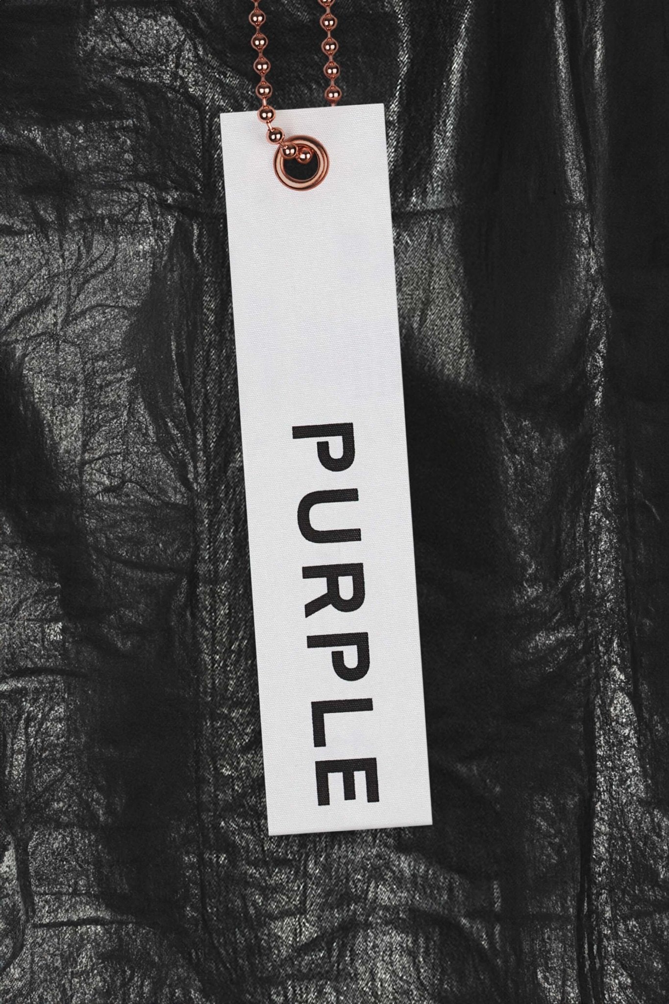 A tag displaying "PURPLE BRAND" in white contrasts against a sleek metallic black backdrop, evoking the edgy allure of Purple Brand P004-CFDB Coated Flare Black denim.