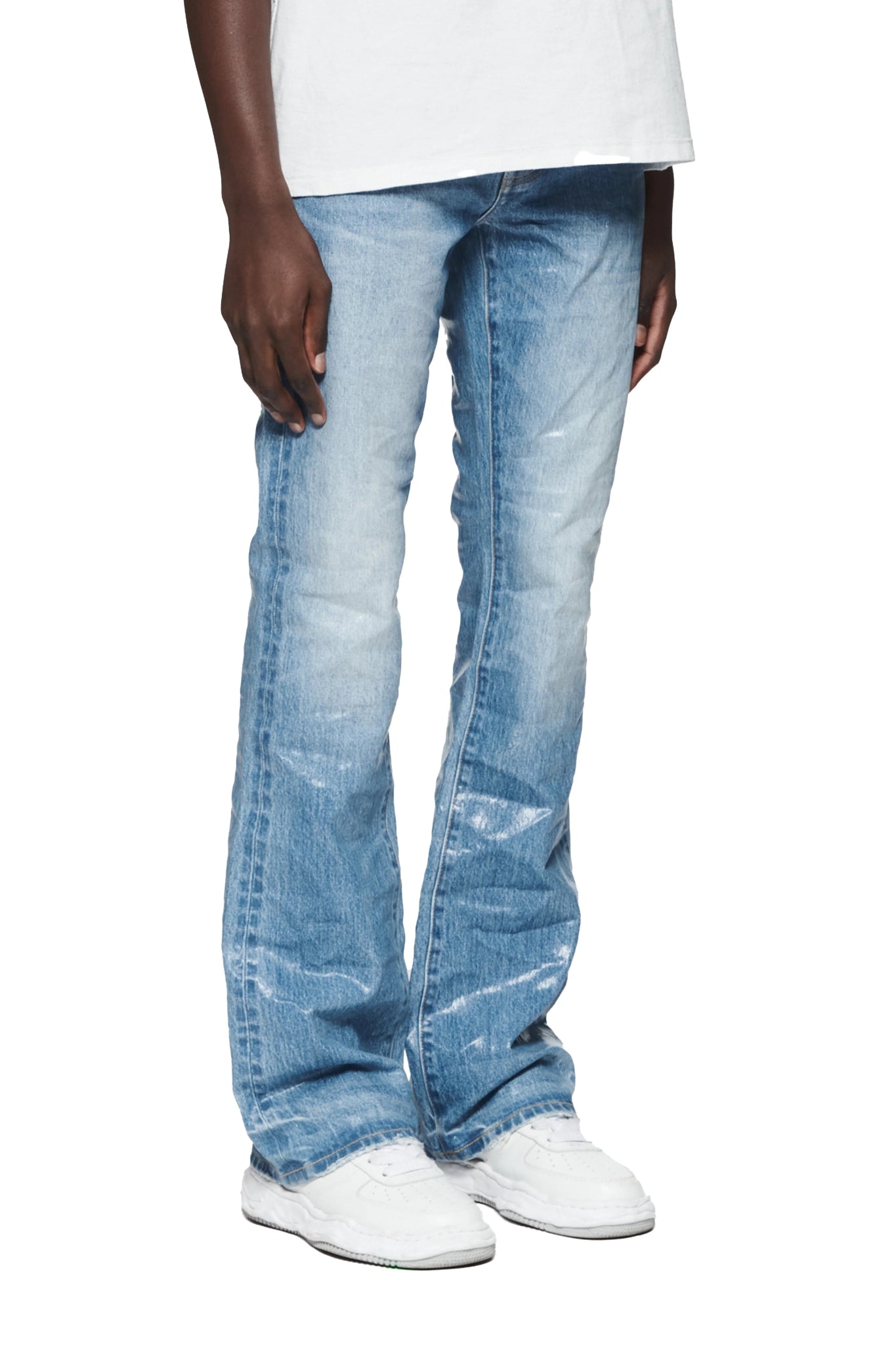 A person wearing PURPLE BRAND P004-CFLC Coated Flare Low Rise Jeans in Light Indigo and white sneakers, with only their legs and lower torso visible.