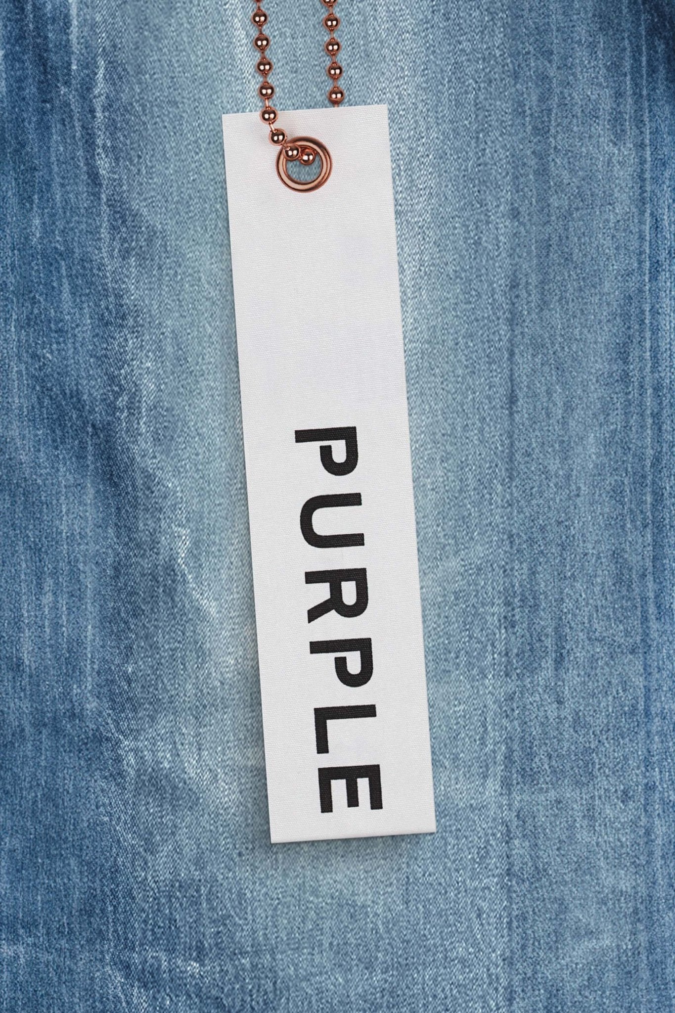 A white tag with the word "PURPLE" in black letters hangs on a ball chain against a backdrop of PURPLE BRAND P004-CFLC COATED FLARE LT INDIGO premium stretch denim.