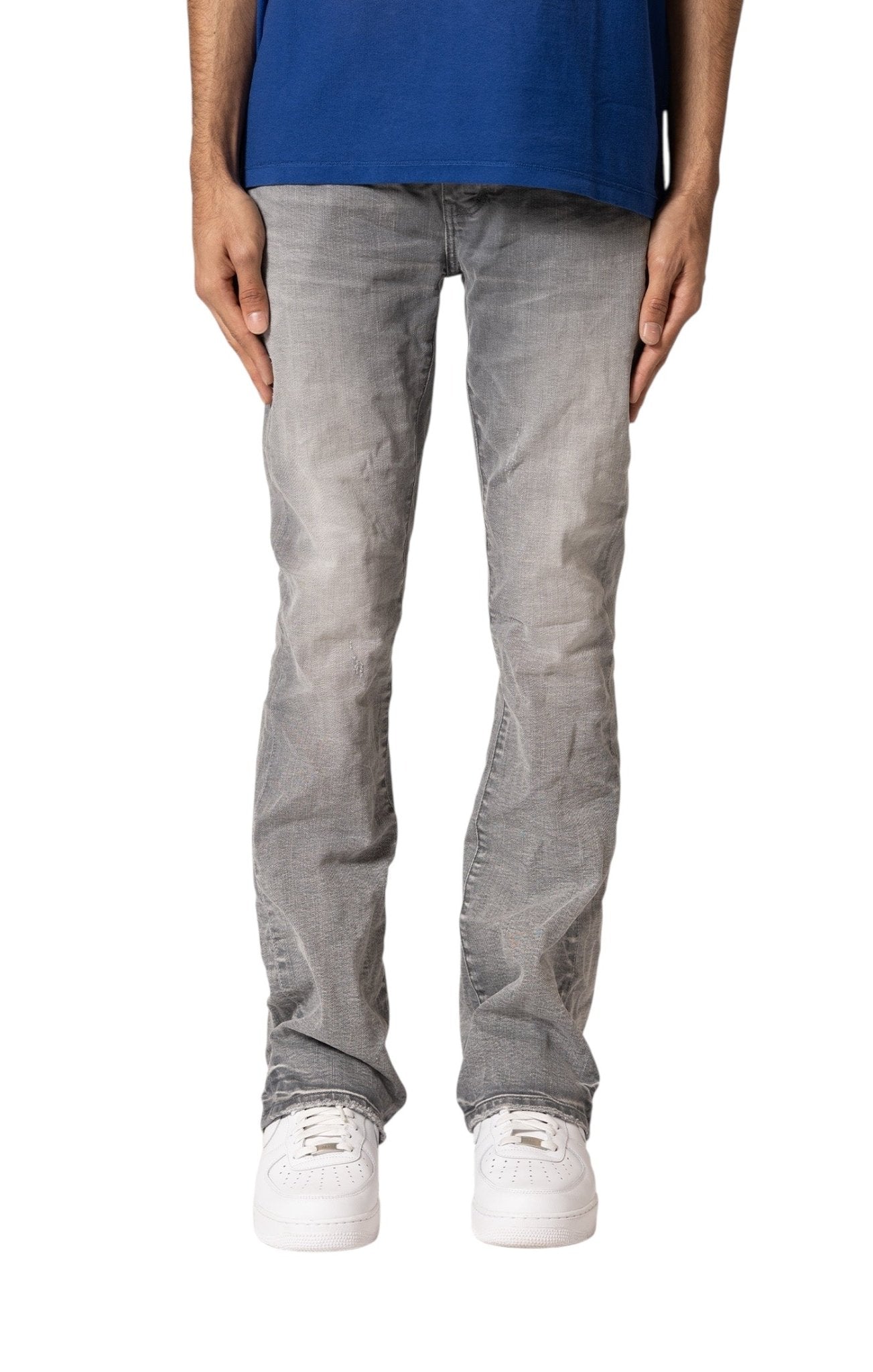 A person wears a blue shirt, white sneakers, and PURPLE BRAND Purple Brand P004-FTMY325 Flare Stormy Ski Grey jeans with a button-fly closure, standing with arms relaxed at their sides.