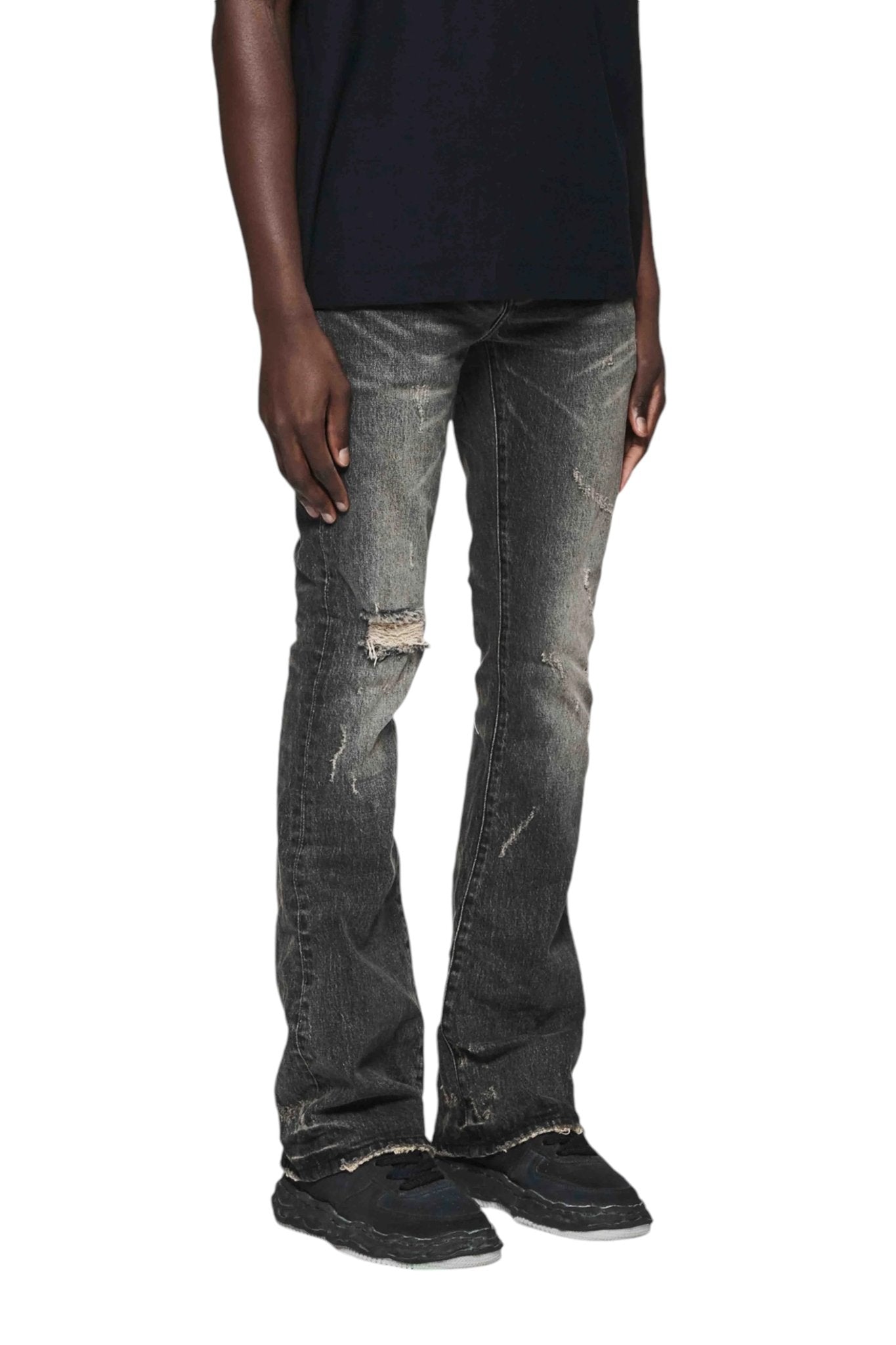 A person is wearing the Purple Brand P004-VFLB Vintage Flare Blowout Black jeans from PURPLE BRAND and black sneakers, standing against a white background.