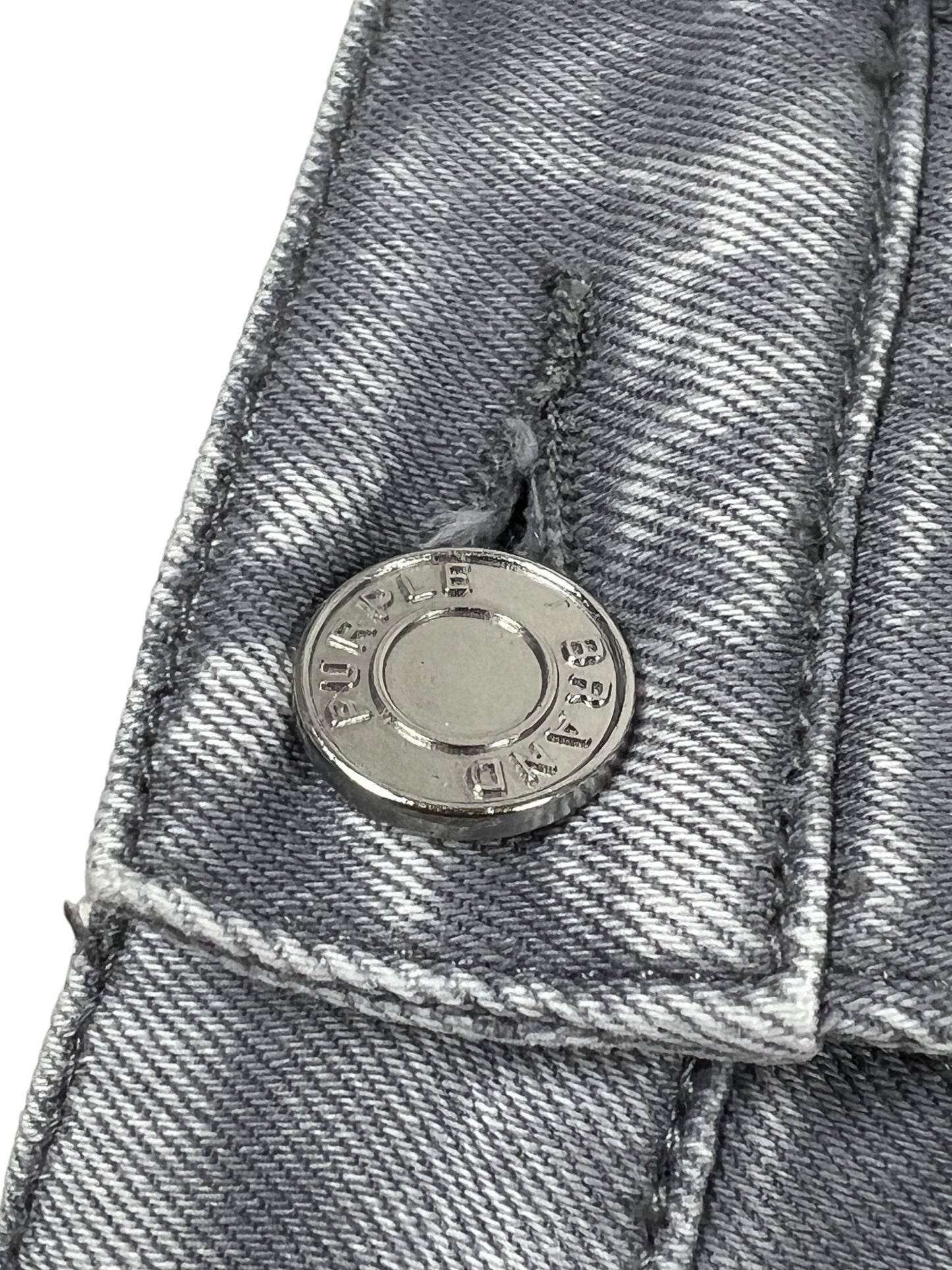 Close-up of a metallic button-fly on Purple Brand P005-AYDG325 Alloy Studs Overdyed Grey jeans, featuring "PURPLE" and numbers engraved—showcasing the fine detail of these premium stretch denim men’s jeans by PURPLE BRAND.