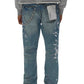 A person in PURPLE BRAND's P005-MGPI Manganese Paint Emb Indigo distressed blue men's jeans, featuring a medium indigo wash with white paint splatters and a keychain on the back pocket, wears a gray hoodie. The premium stretch denim leads to crisp white sneakers visible on their feet.