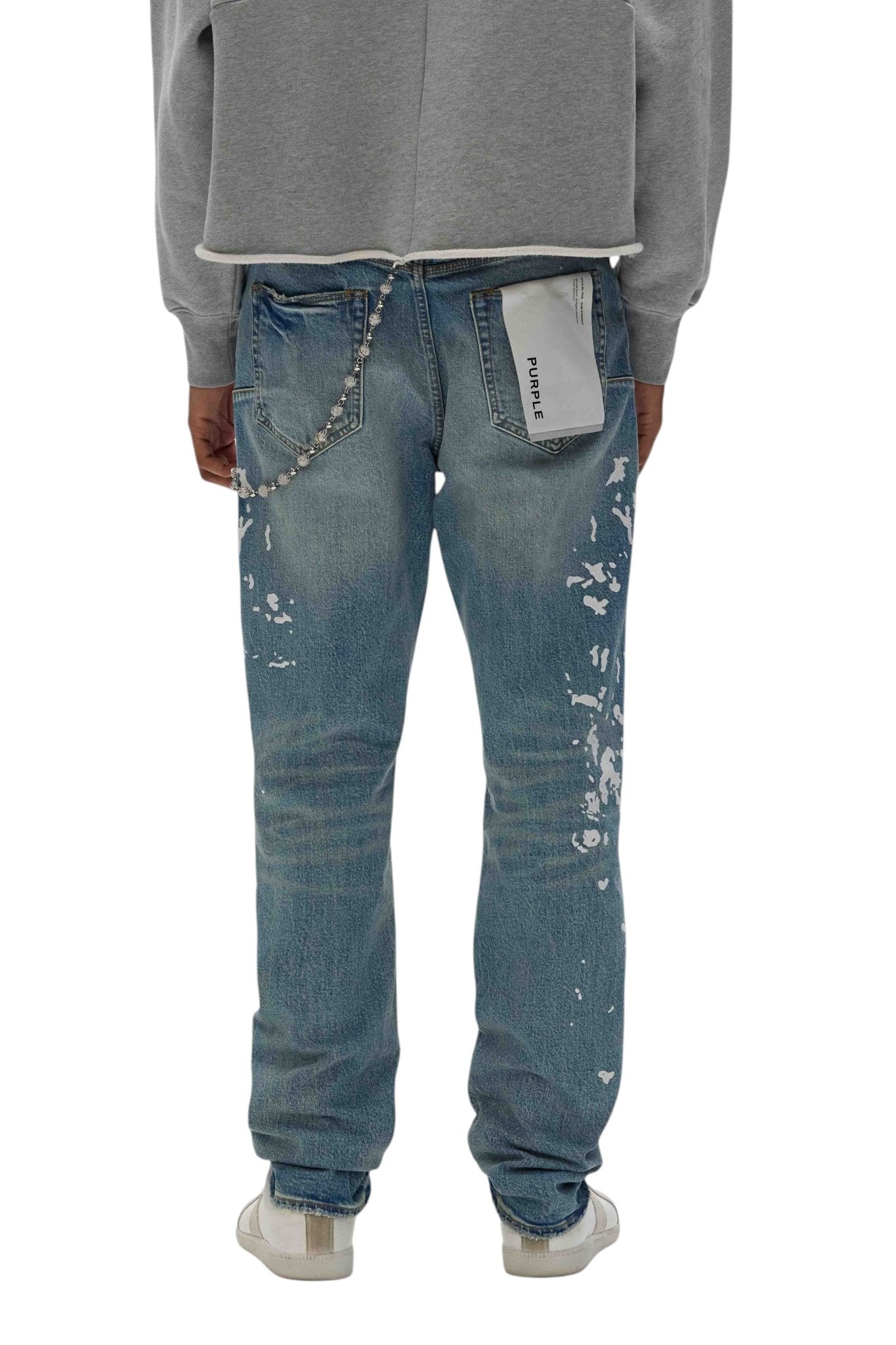 A person in PURPLE BRAND's P005-MGPI Manganese Paint Emb Indigo distressed blue men's jeans, featuring a medium indigo wash with white paint splatters and a keychain on the back pocket, wears a gray hoodie. The premium stretch denim leads to crisp white sneakers visible on their feet.